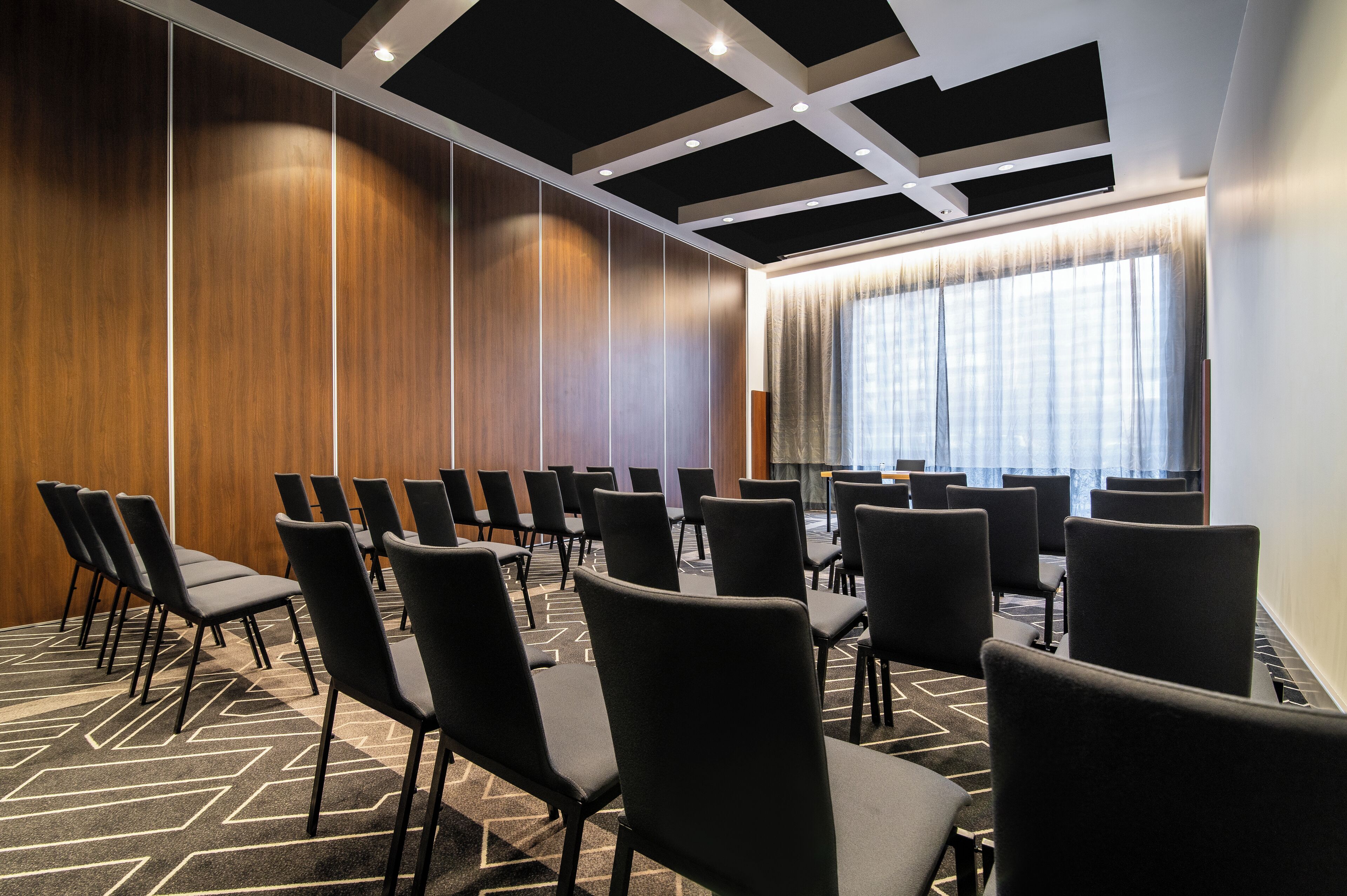 Un salón de conferencias con filas de sillas negras, alfombra estampada, paneles de madera y un ventanal con cortinas.