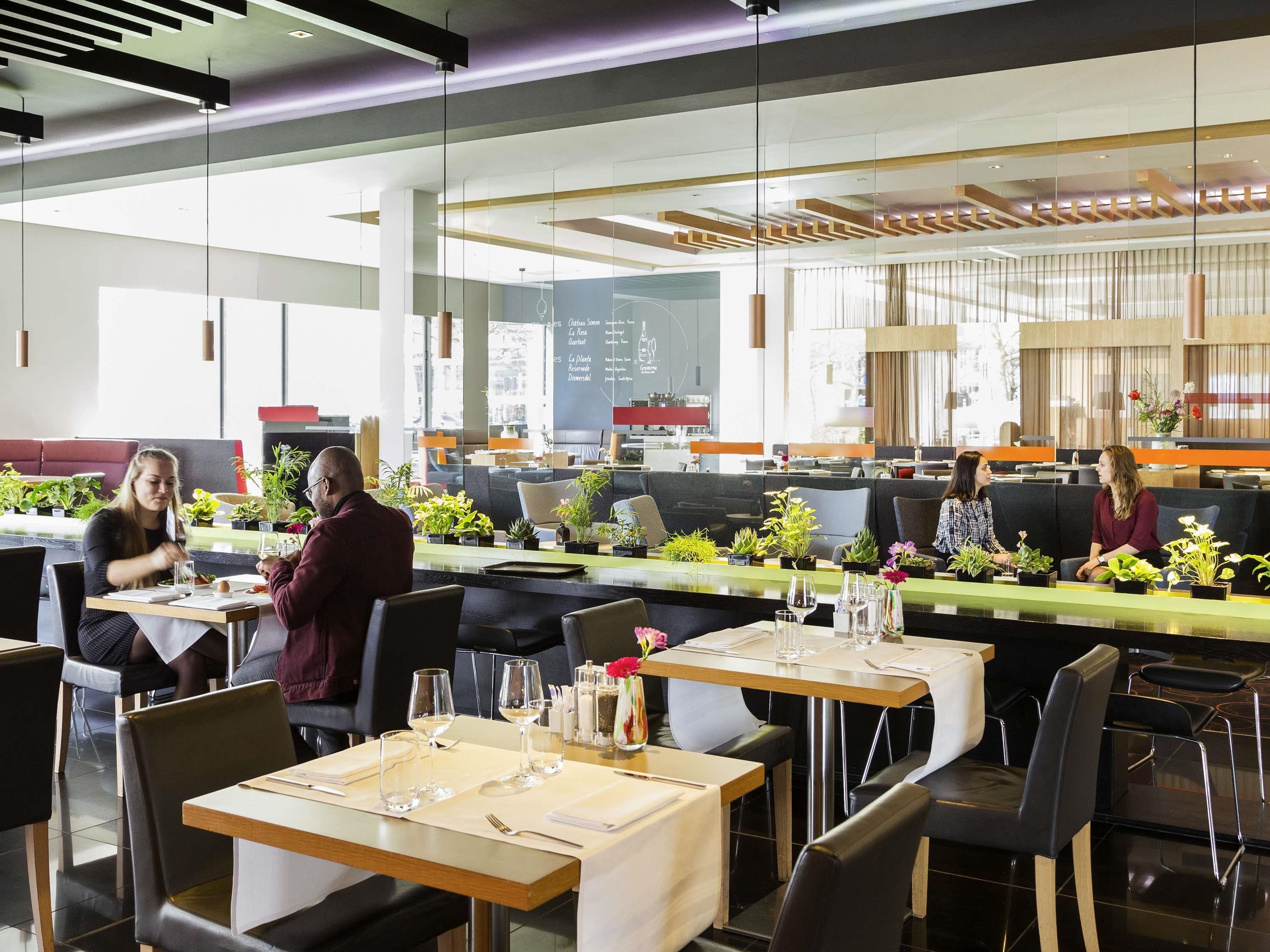 Un restaurante moderno con separadores de vidrio, mesas dispuestas para comer y diversas plantas sobre ellas.