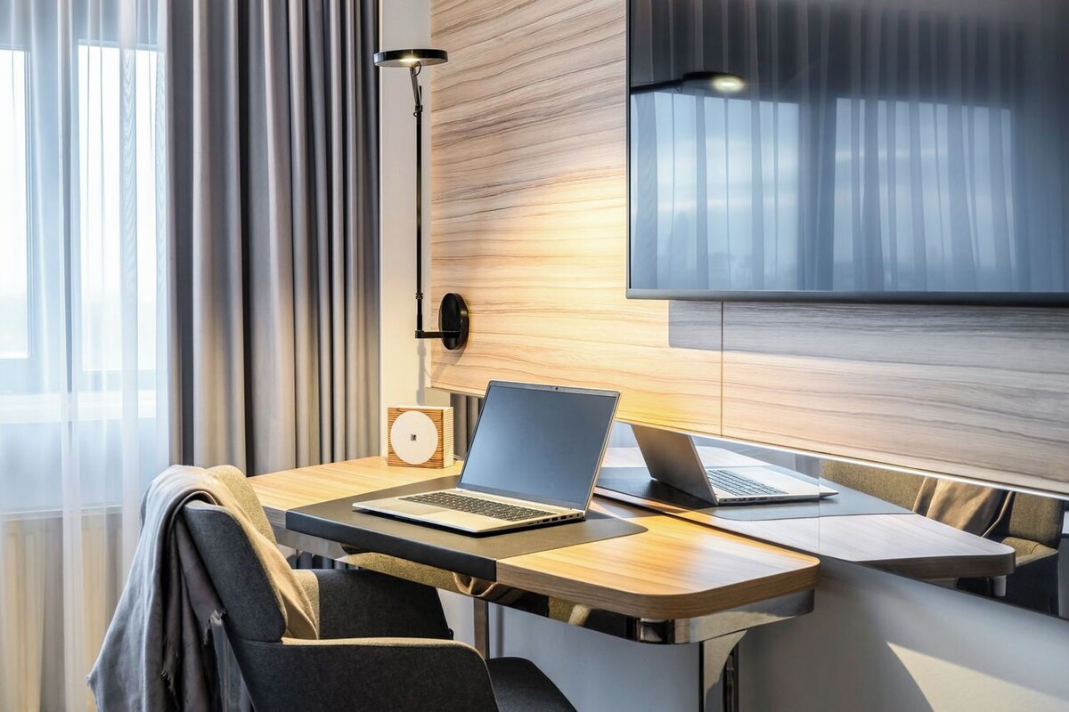 Ein modernes Hotelzimmer mit einem Holztisch, zwei Laptops, einer Uhr und einer Lampe.