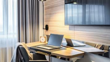 A modern hotel room with a wooden desk, two laptops, a clock, and a lamp.
