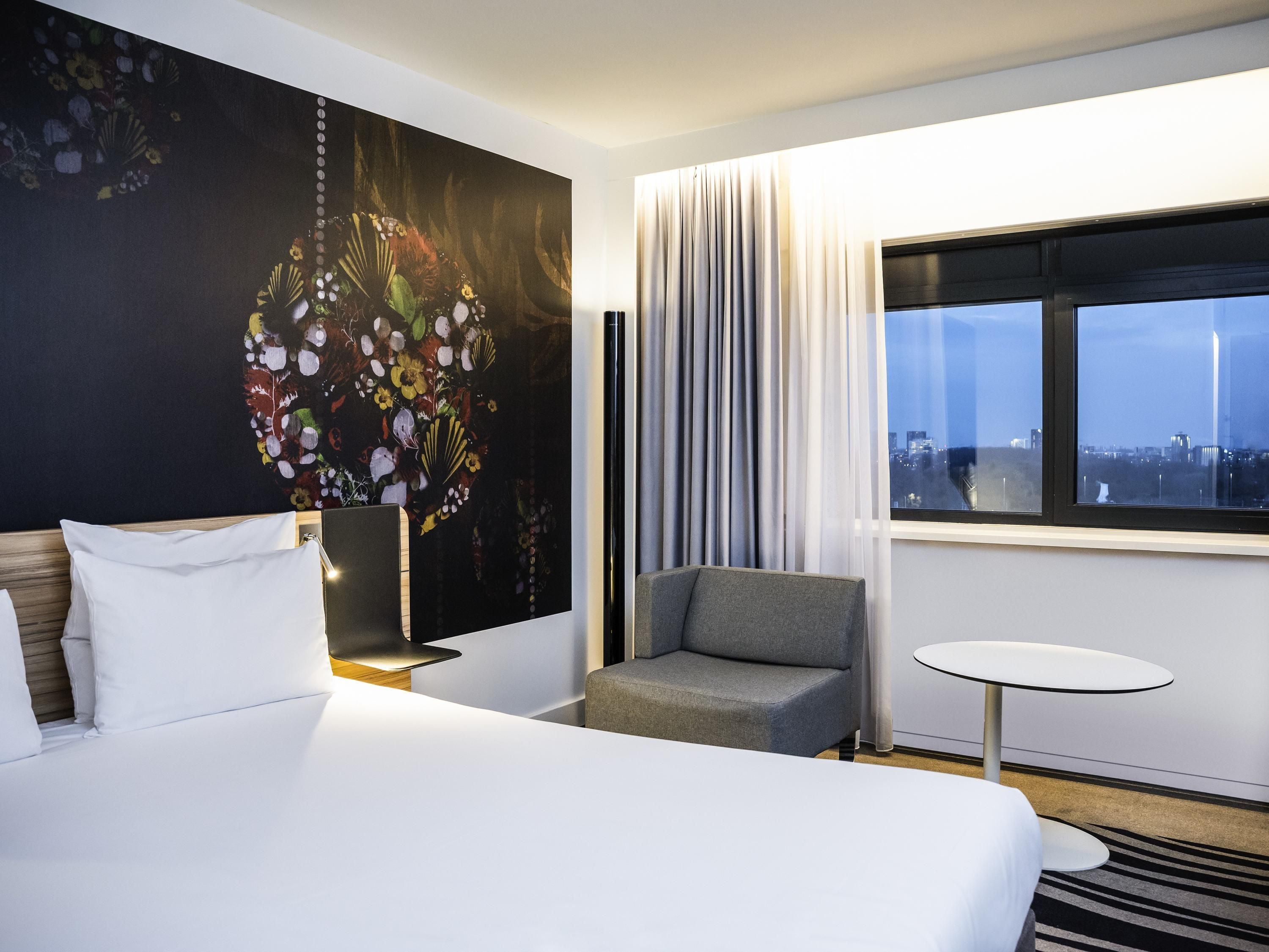 Quarto de hotel com uma cama grande, uma poltrona cinzenta, uma mesinha redonda e vista para a cidade através de uma janela.
