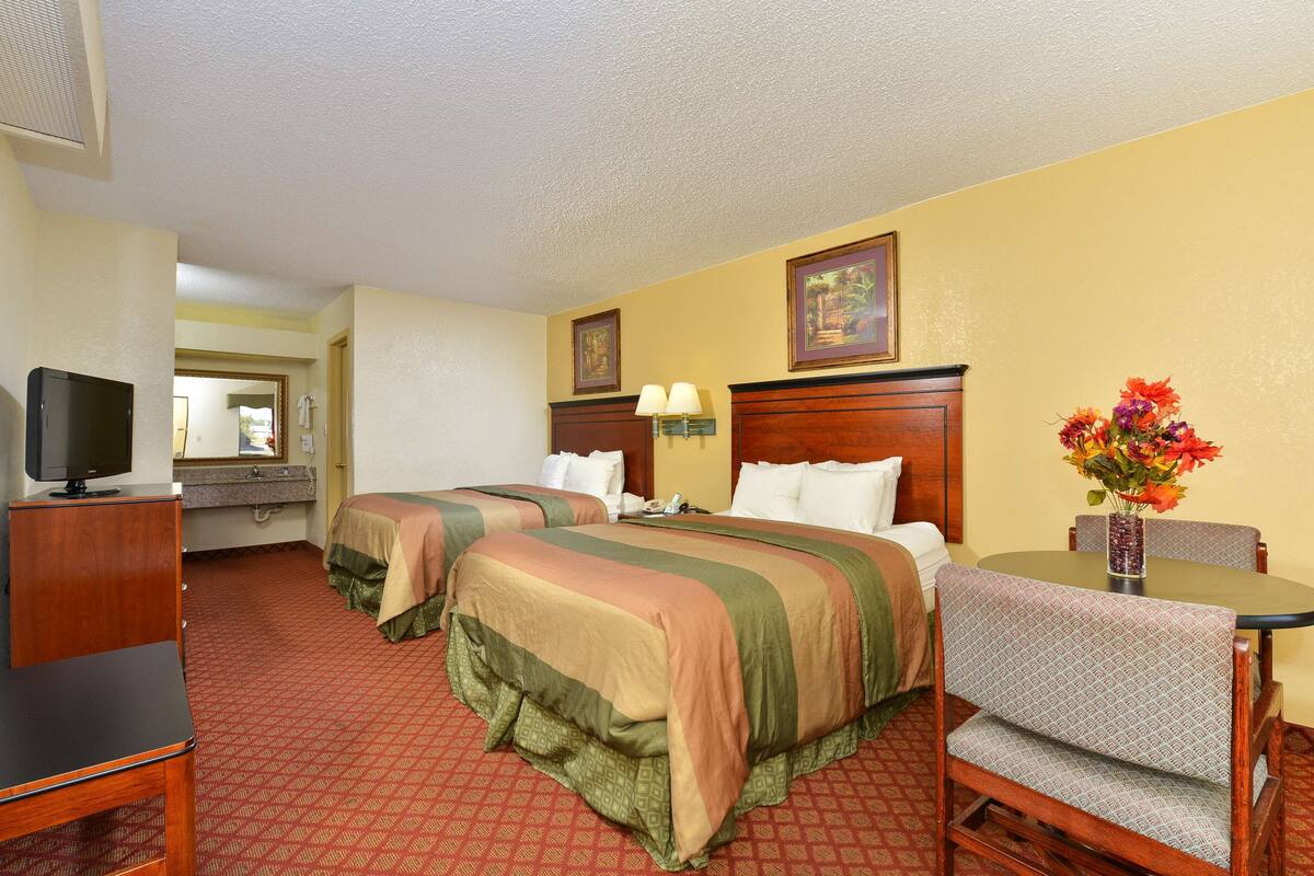 A hotel room with two beds, a TV, a desk with a chair, and a vase with flowers.