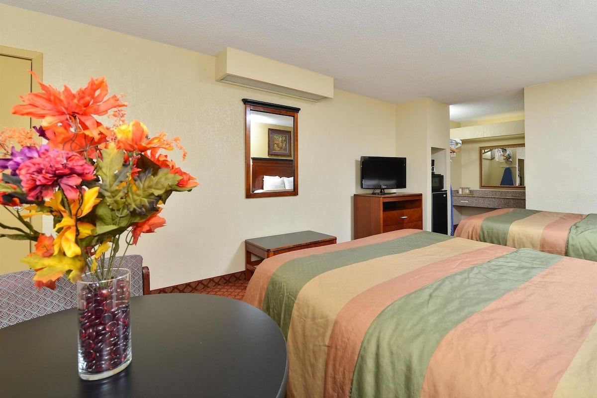 A hotel room with two beds, a television, a small table with a vase of flowers, and a mirror.