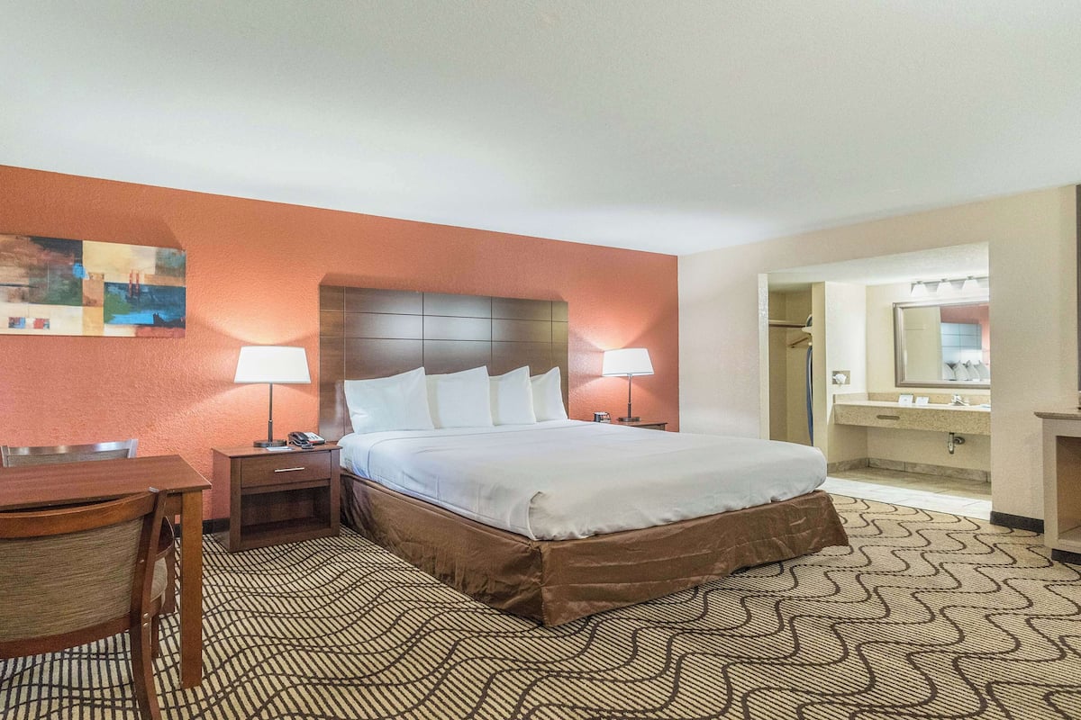 A hotel room with a bed, bedside tables, a desk, a chair, a bathroom with a mirror, and a patterned carpet.