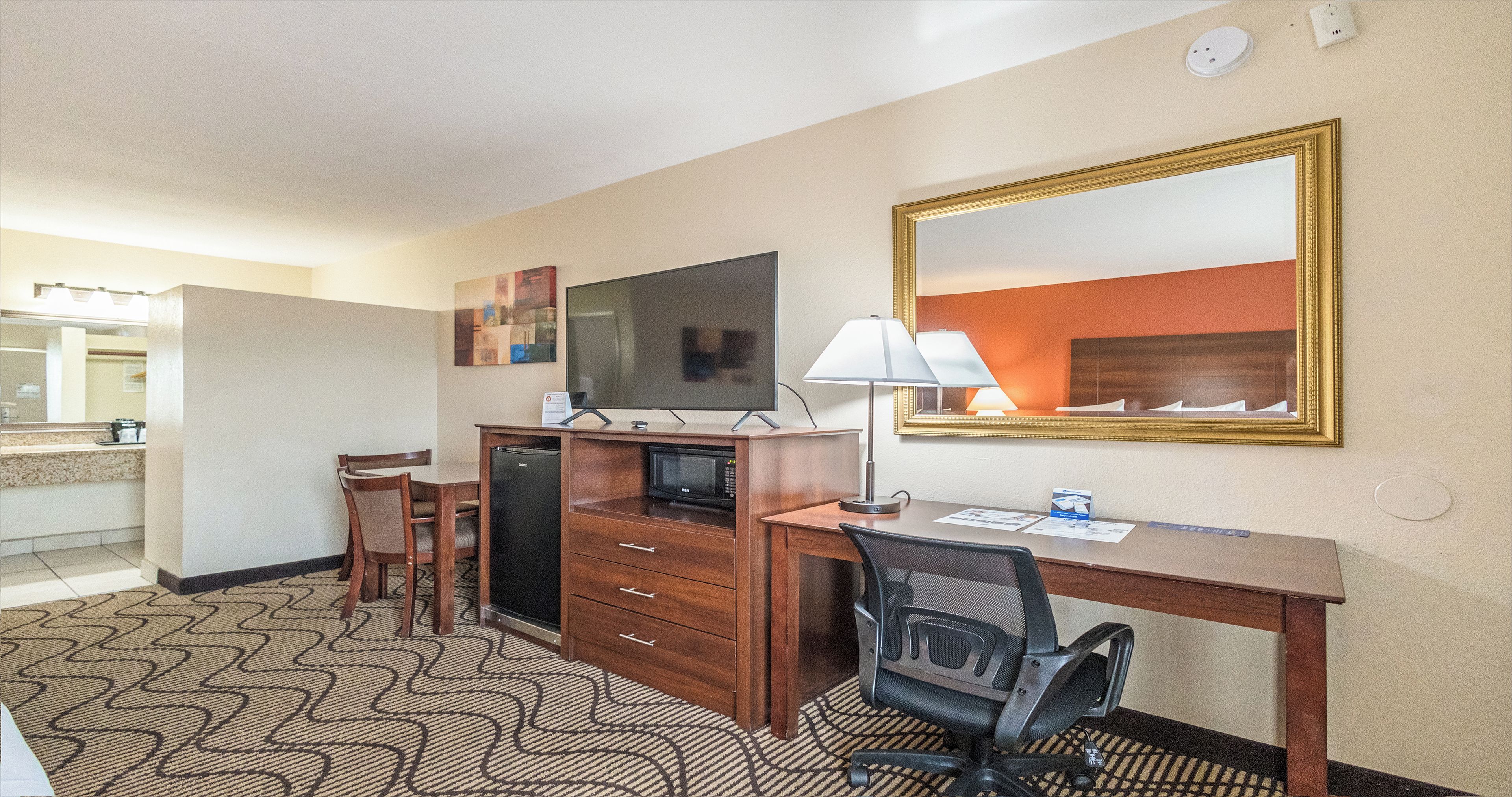 A hotel room with a desk, chair, television, microwave, and a large mirror.