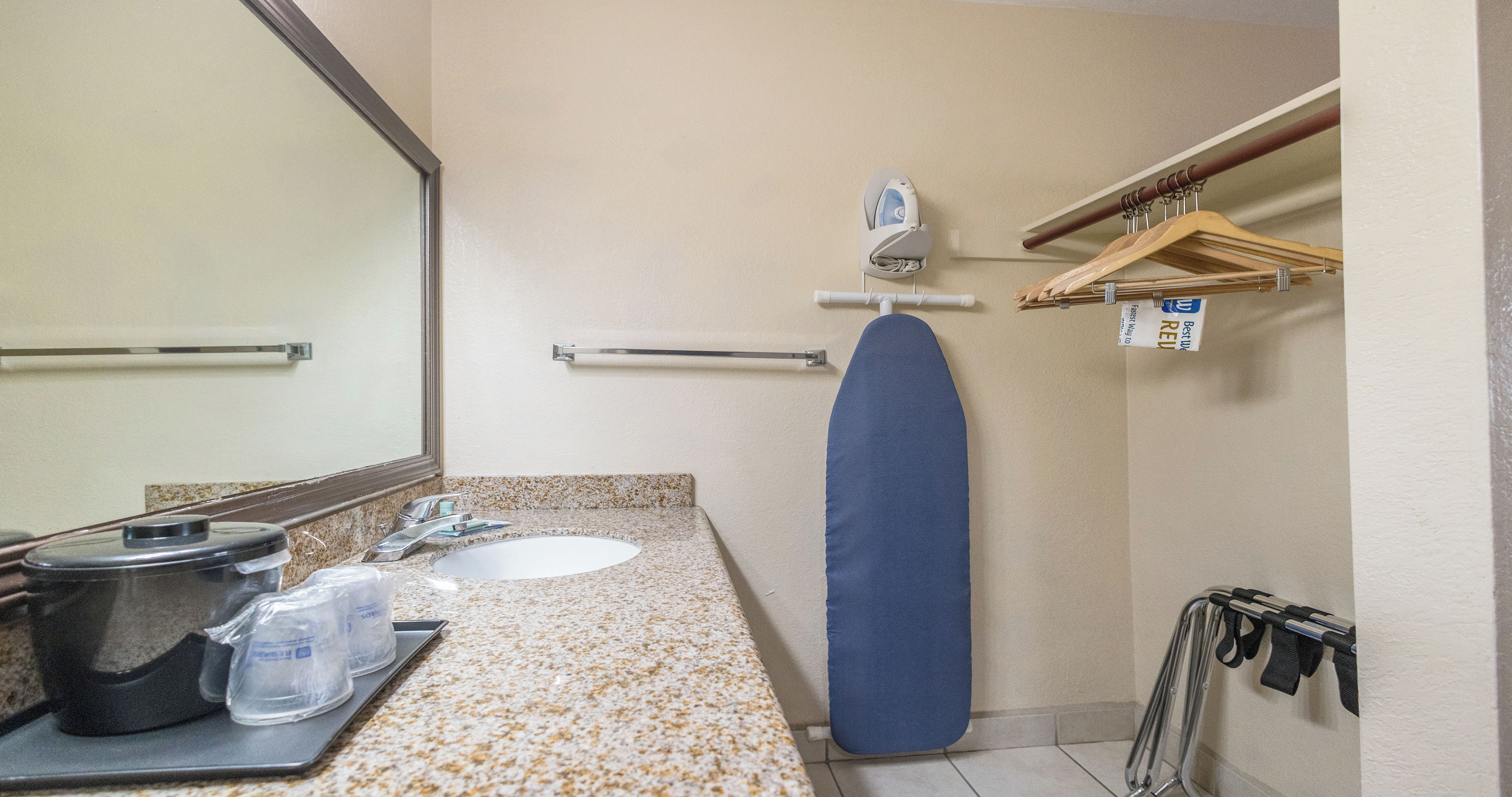 A bathroom with a sink, mirror, towel rack, ironing board, and a shower area.