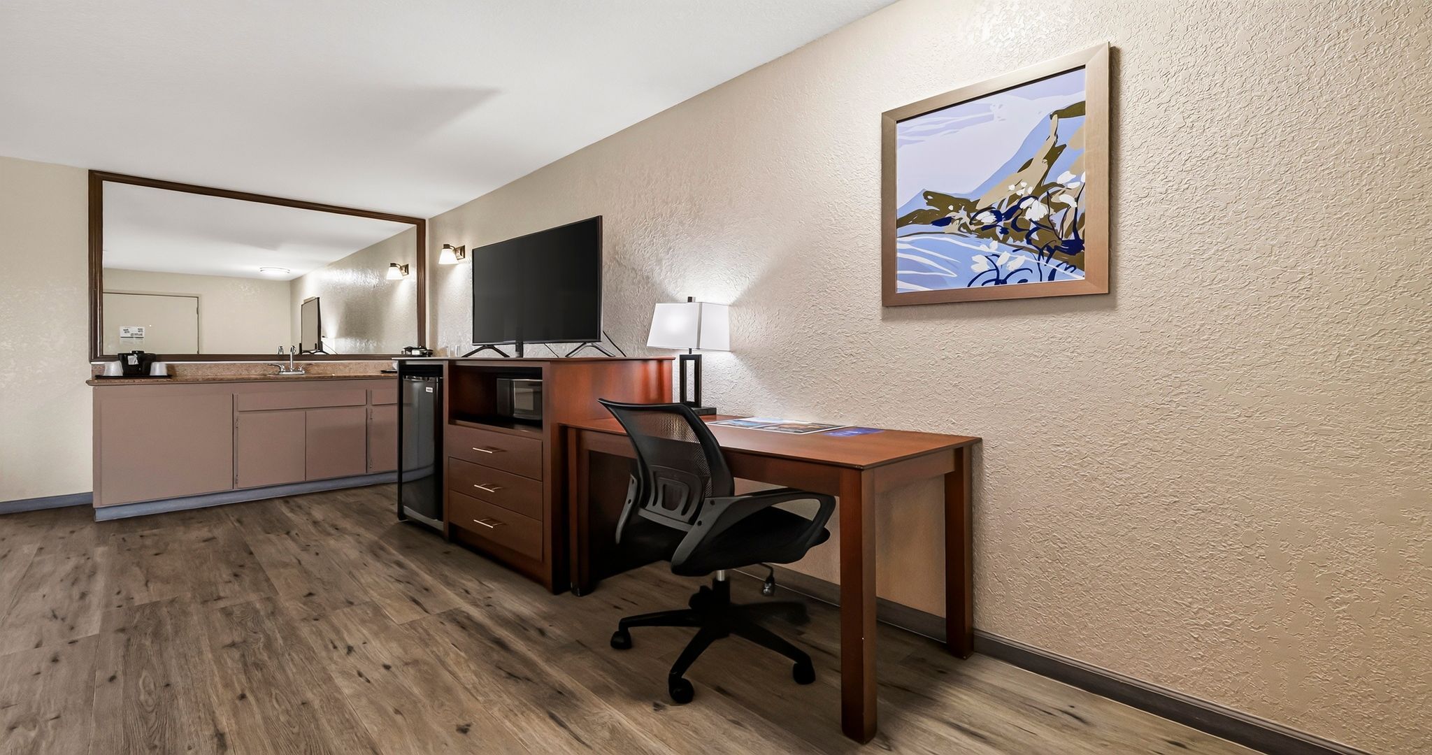 A hotel room with a wooden desk, a chair, a television, a small kitchenette, and a painting on the wall.