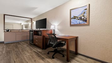 A hotel room with a wooden desk, a chair, a television, a small kitchenette, and a painting on the wall.