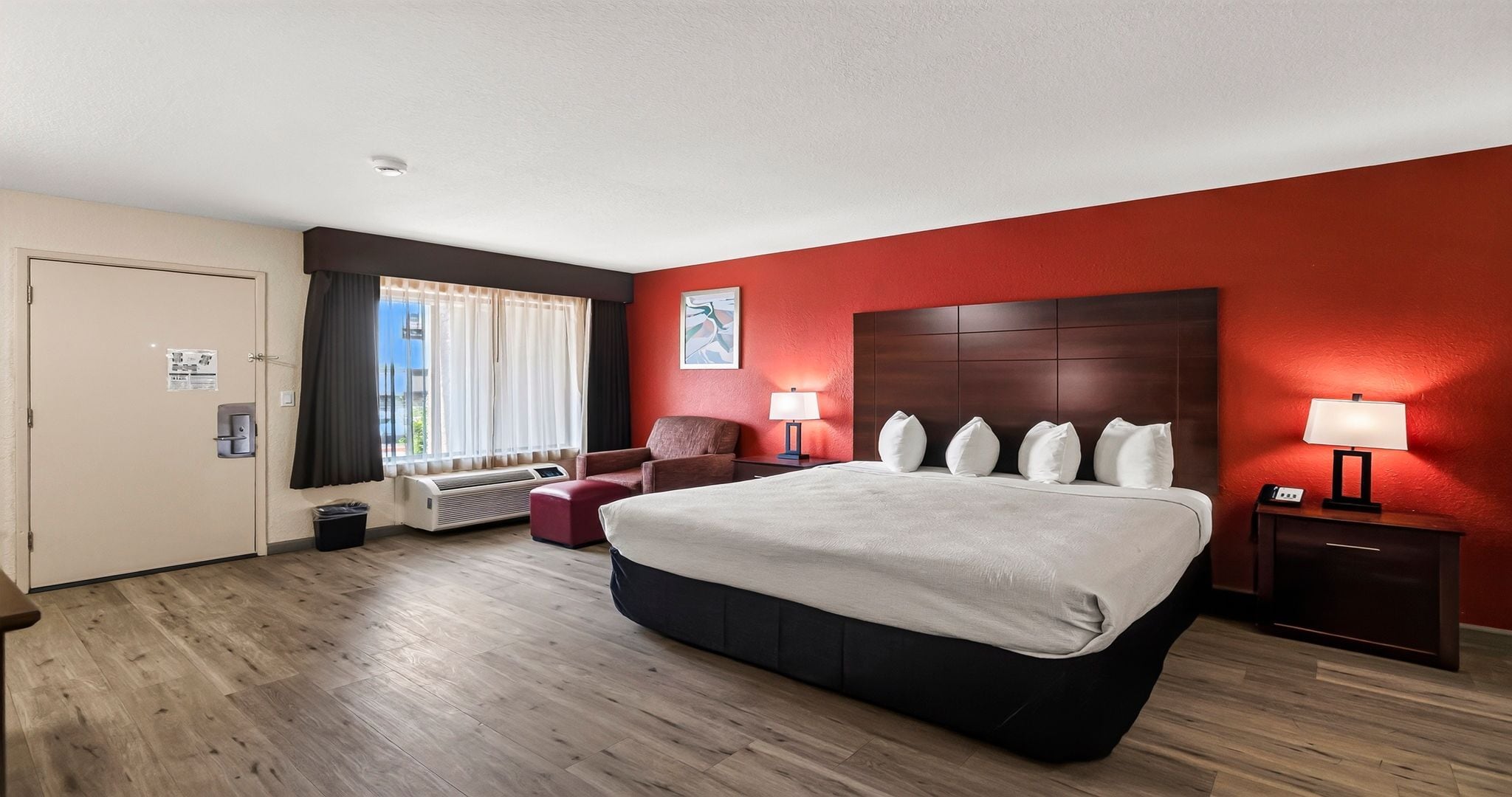 A hotel room with a large bed, a chair, a bedside table with a lamp, a window with curtains, and a door.