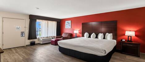 Habitación de hotel con una cama grande, una silla, una mesita de noche con lámpara, una ventana con cortinas y una puerta.