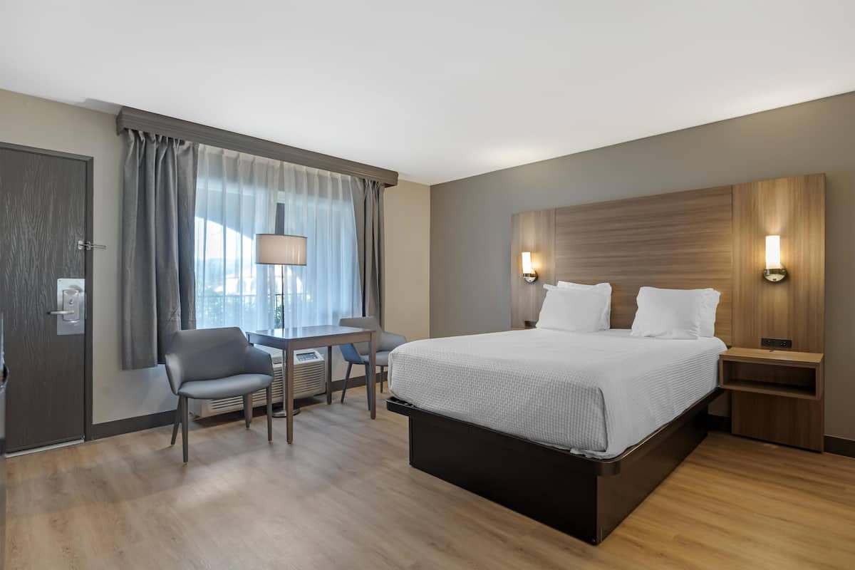 A hotel room with a bed, desk, chair, and a view of the outdoors through the window.