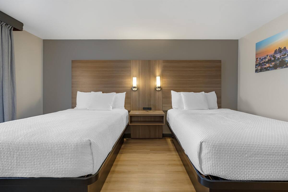 A hotel room with two single beds, a wooden headboard, two wall-mounted lights, a nightstand, and a cityscape painting on the wall.