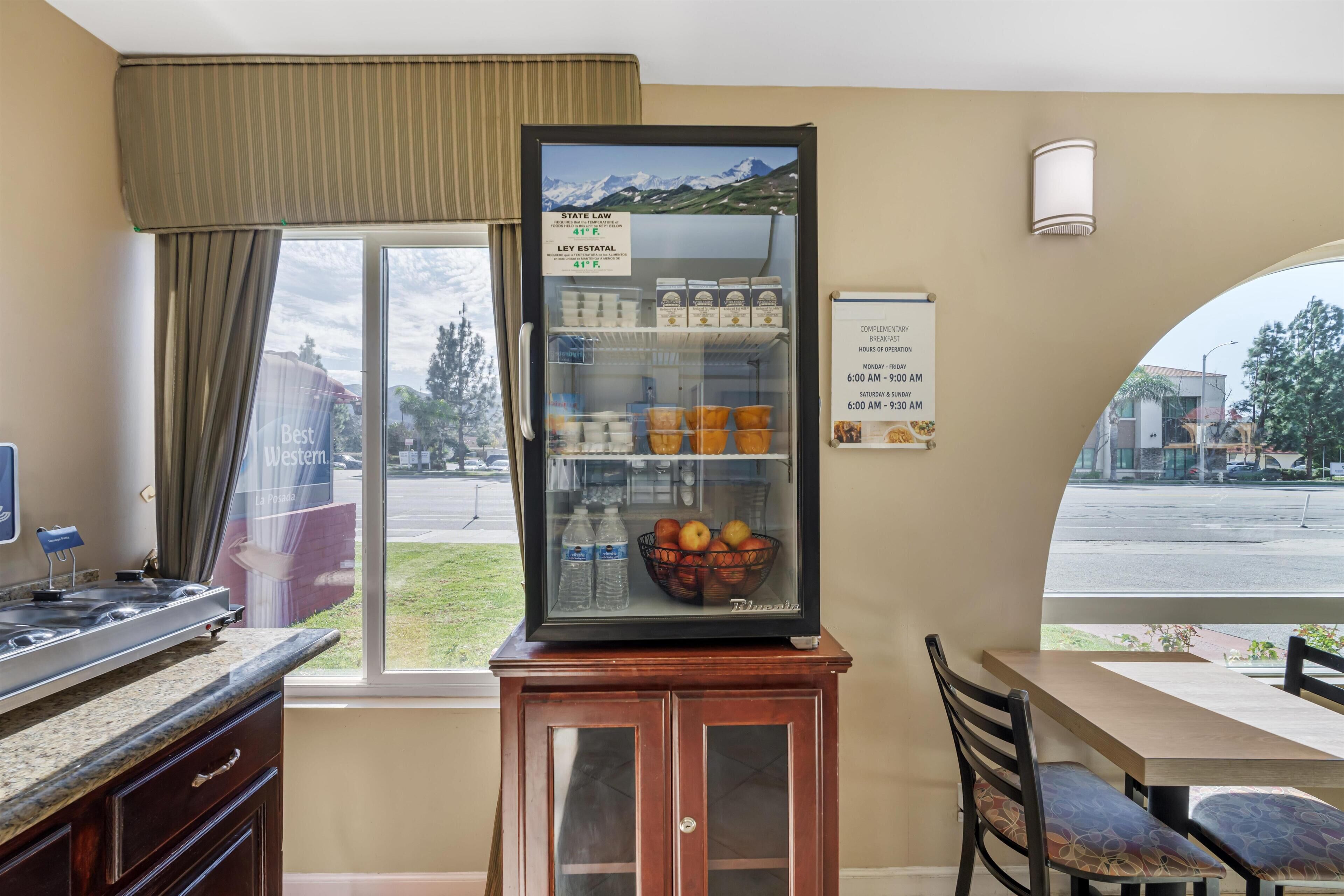 Una cocina con un refrigerador lleno de bebidas y frutas, una zona de comedor con mesa y sillas, y vista al exterior.