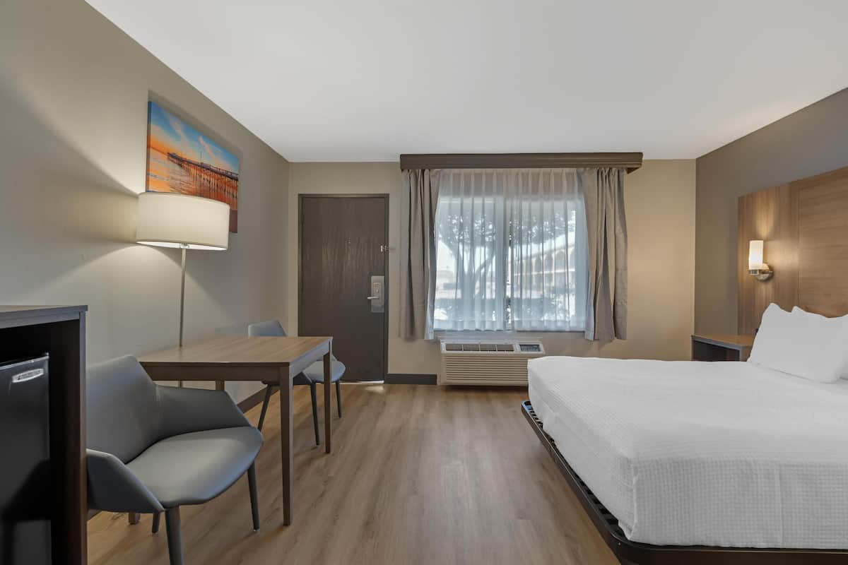 A hotel room with a bed, a desk, a chair, a TV, a painting, a window with curtains, and a wall-mounted lamp.