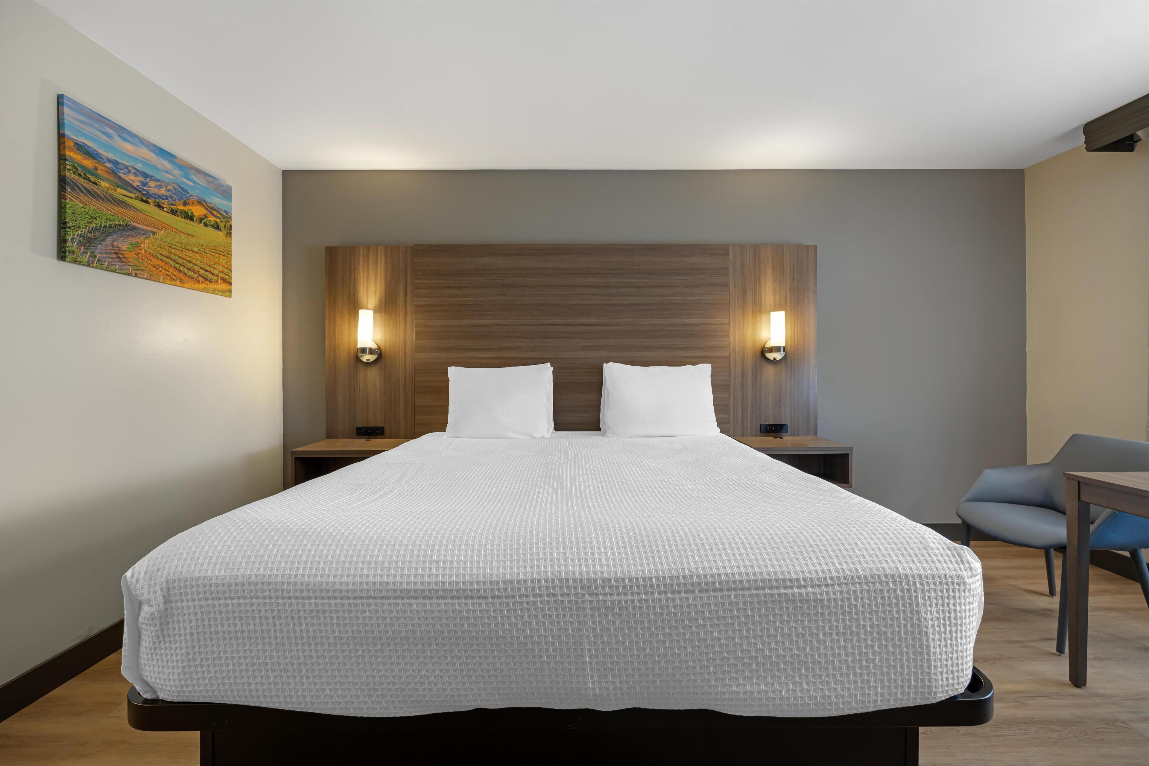 Une chambre d’hôtel moderne avec un grand lit, deux tables de chevet, une petite table, une chaise et un tableau au mur.