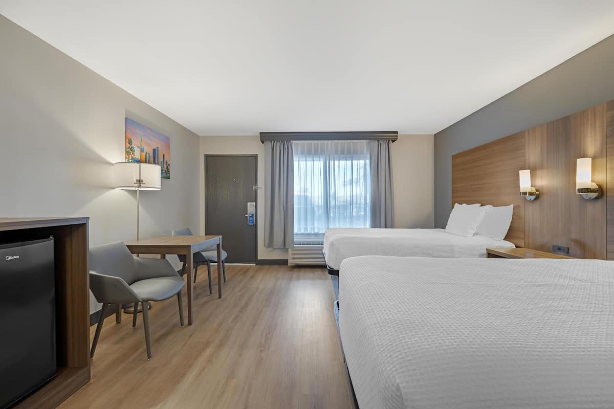 A hotel room with two beds, a desk, a chair, a refrigerator, and a painting on the wall.