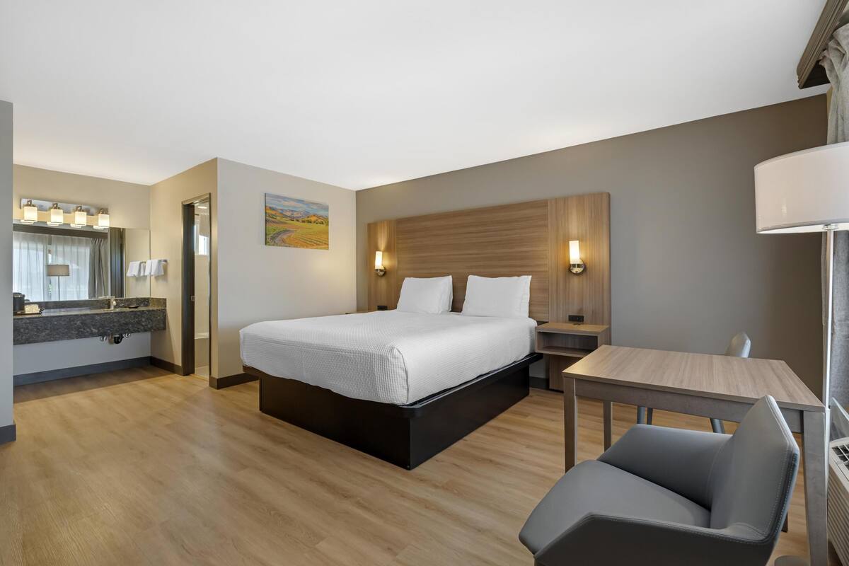 A modern hotel room with a bed, bedside table, chair, and a small desk with a lamp.