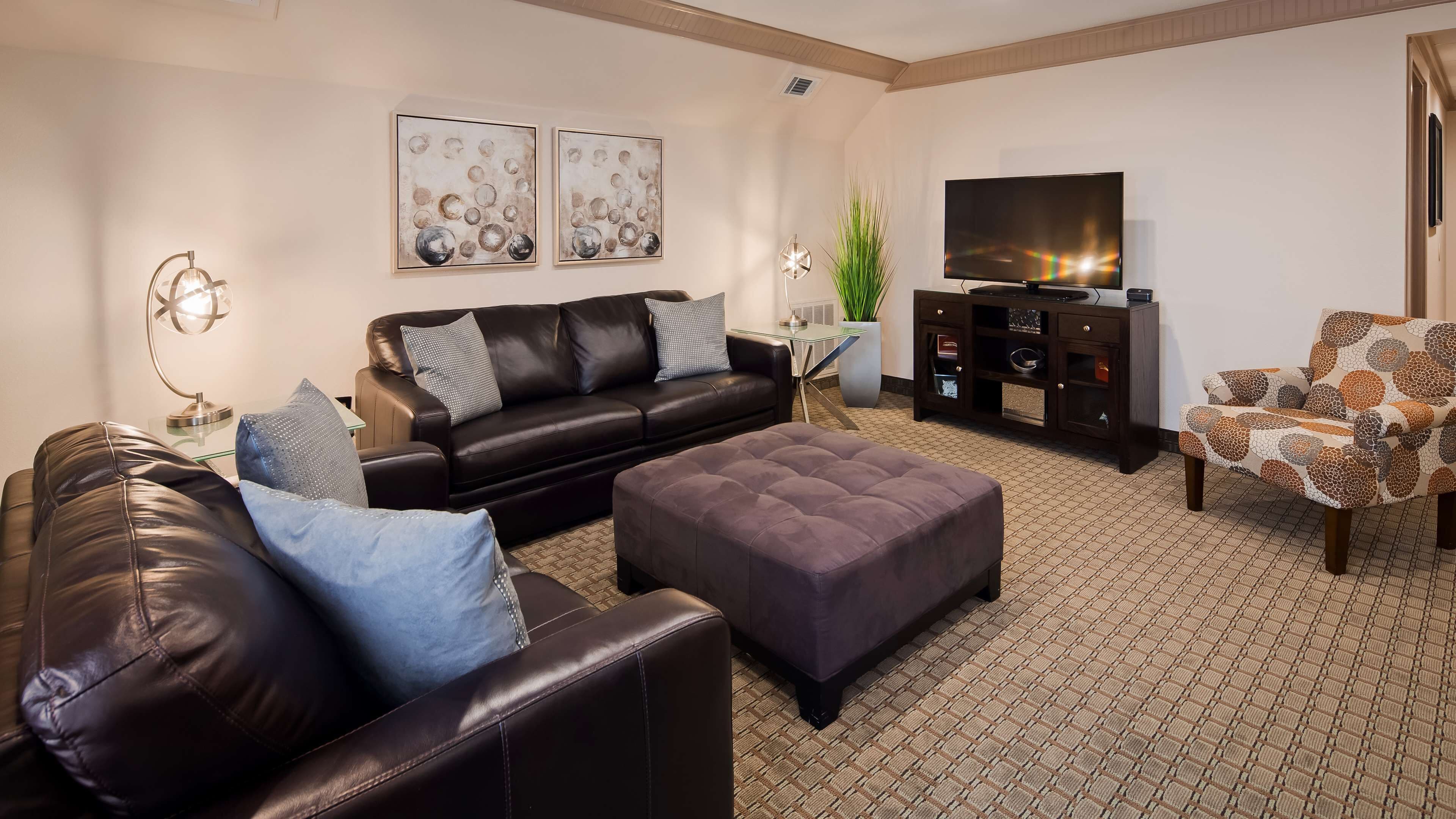 Ein modernes Wohnzimmer mit Ledersofa, Hocker und gemustertem Sessel. An der Wand befinden sich ein Flachbildfernseher und ein Schrank.