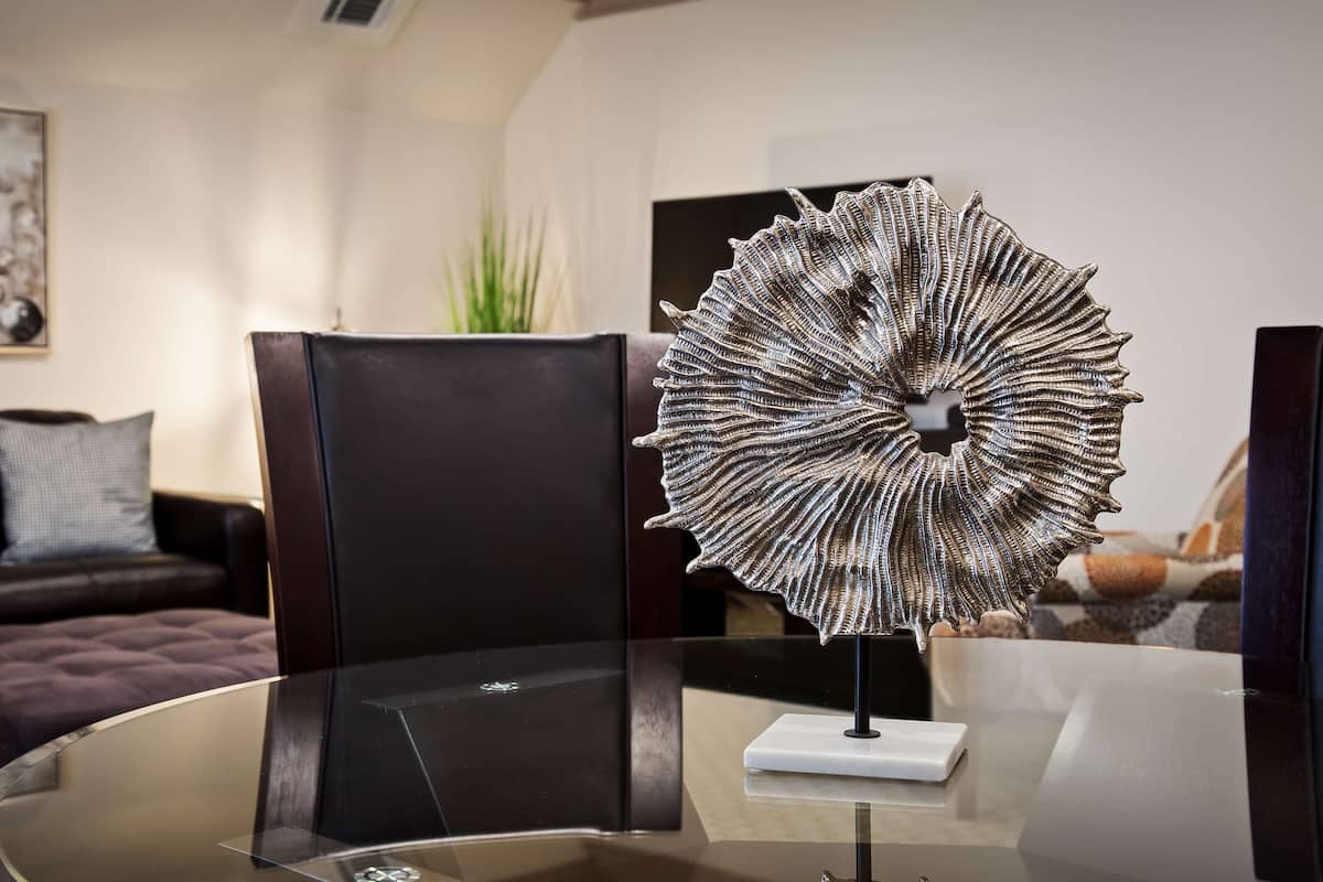 A modern living room with a glass coffee table, a decorative metal sculpture, a dark leather sofa, and a framed abstract painting on the wall.