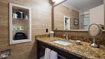 Un baño con encimera de granito, lavamanos, espejo y repisas con artículos de aseo.