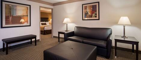 A hotel room with a black leather sofa, a matching ottoman, a small bench, two bedside tables with lamps, and two framed abstract paintings on the walls.