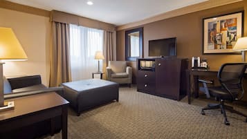 Quarto de hotel com sofá, uma cadeira, escrivaninha, televisão, minibar e um quadro na parede.