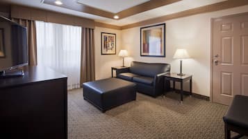A hotel room with a dark leather sofa, a matching ottoman, a desk with a flat-screen TV, two bedside lamps, and two framed artworks on the walls.