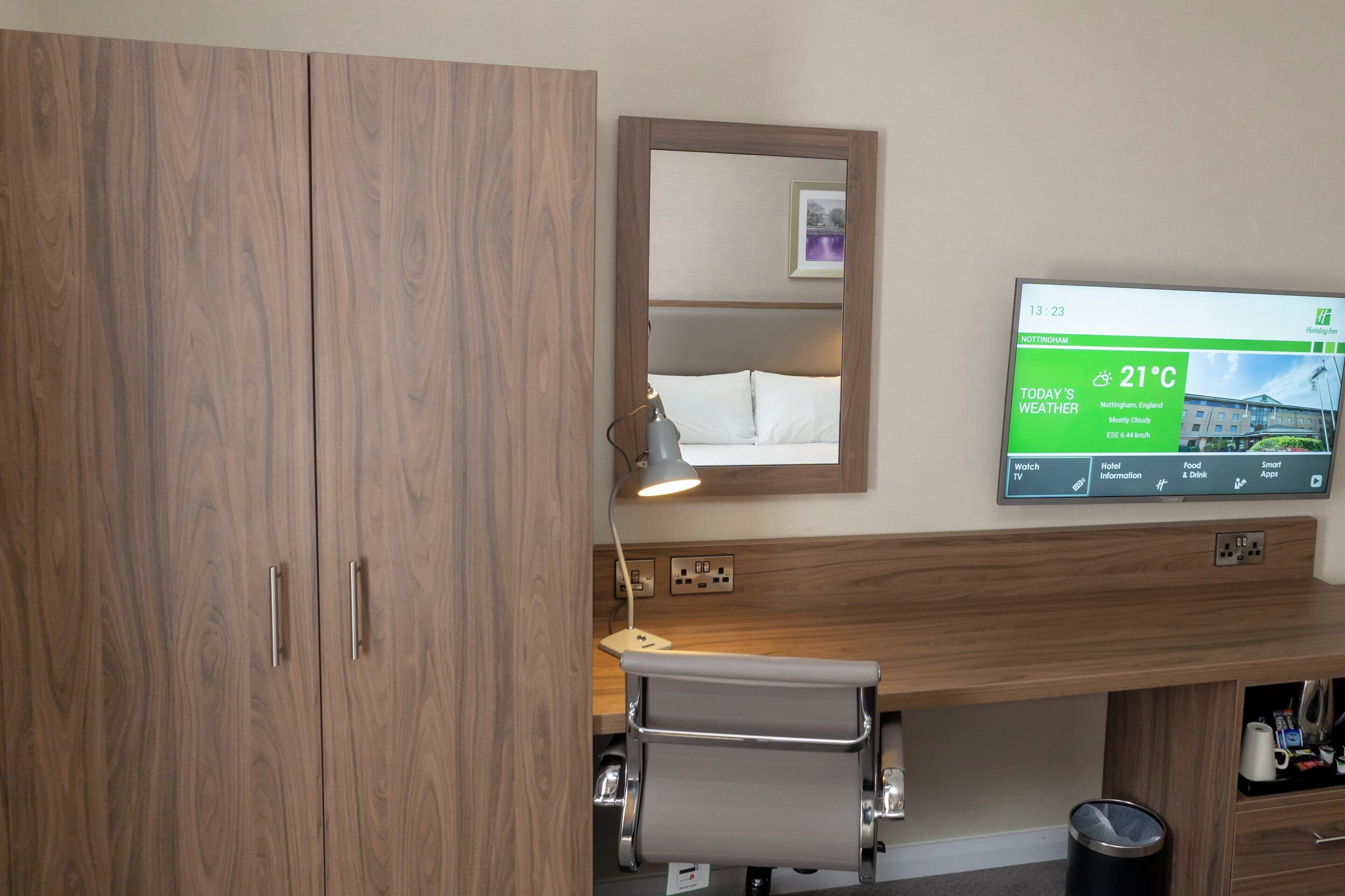 Une chambre d’hôtel avec une armoire en bois, une table de chevet avec une lampe, un téléviseur fixé au mur qui affiche la météo et une petite poubelle.
