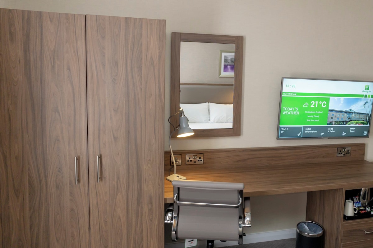 A hotel room with a wooden wardrobe, a bedside table with a lamp, and a TV displaying the weather.