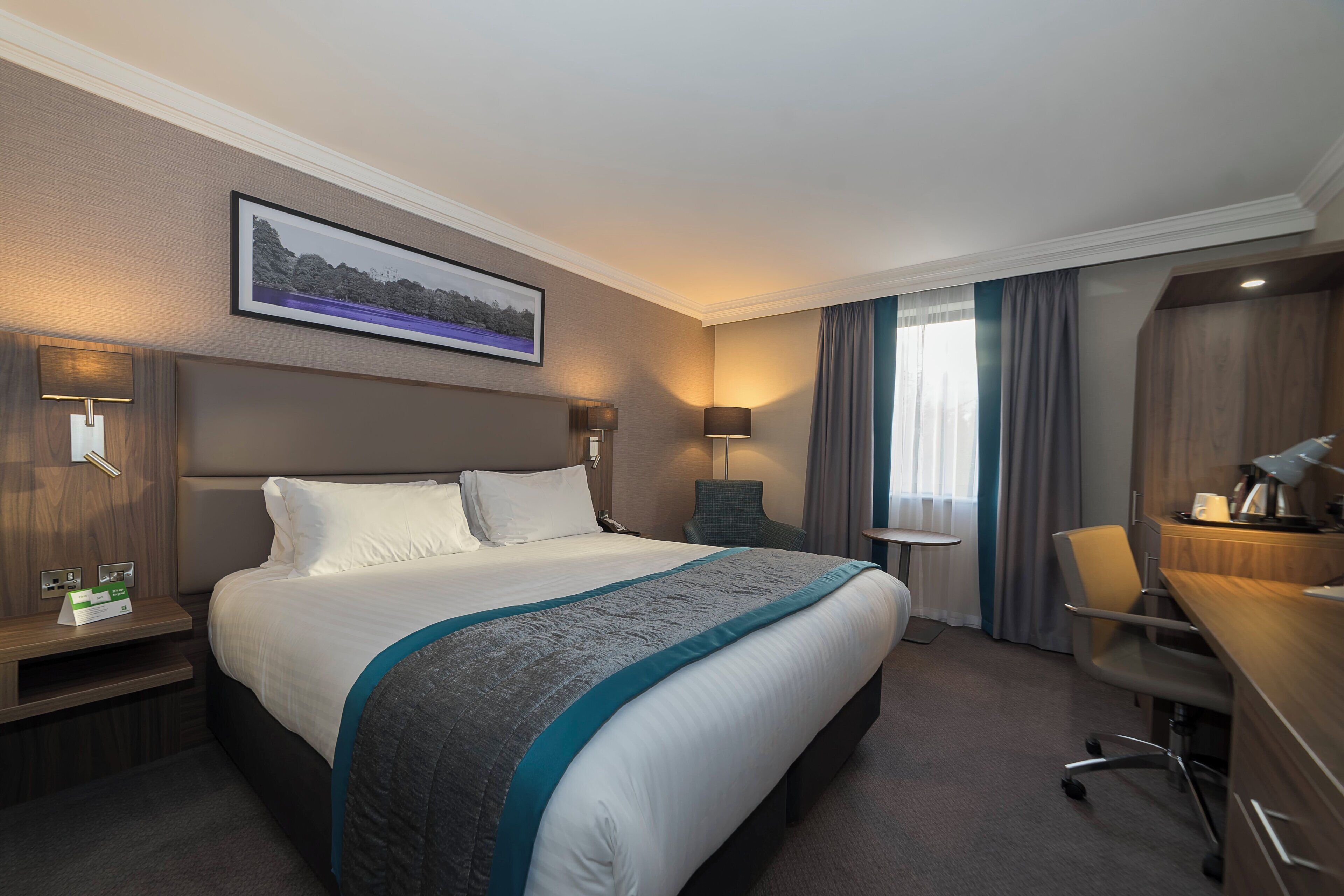 A hotel room with a large bed, a desk, a chair, a lamp, and a painting on the wall.