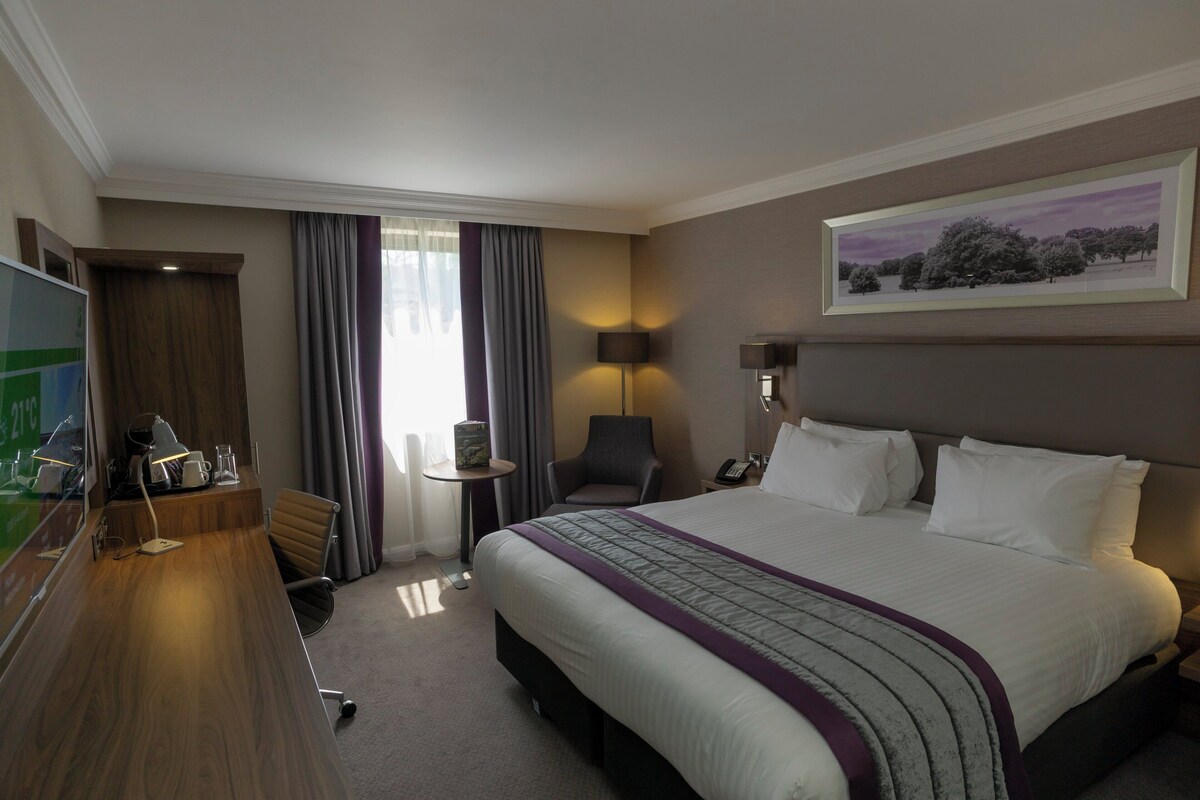 A hotel room with a large bed, a desk with a computer, a chair, a small table, and a lamp.
