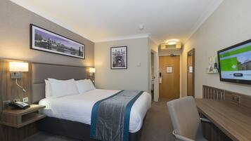 A hotel room with a bed, bedside table with a phone, a desk with a chair, a TV, and framed pictures on the walls.