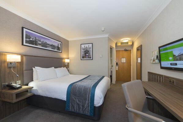 A hotel room with a bed, bedside table with a phone, a desk with a chair, a TV, and framed pictures on the walls.