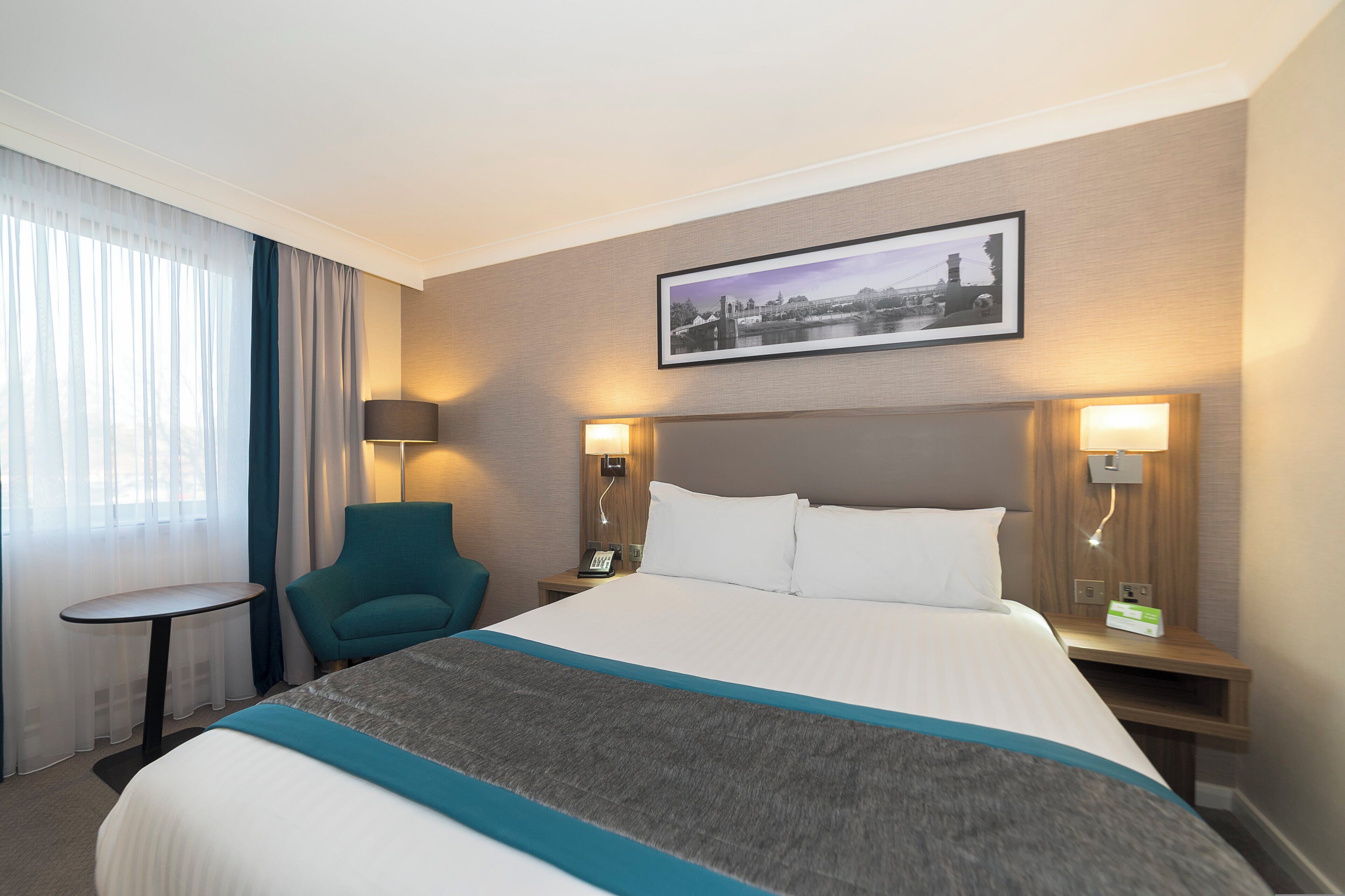 A hotel room with a bed, a chair, a small table, and a wall-mounted picture.