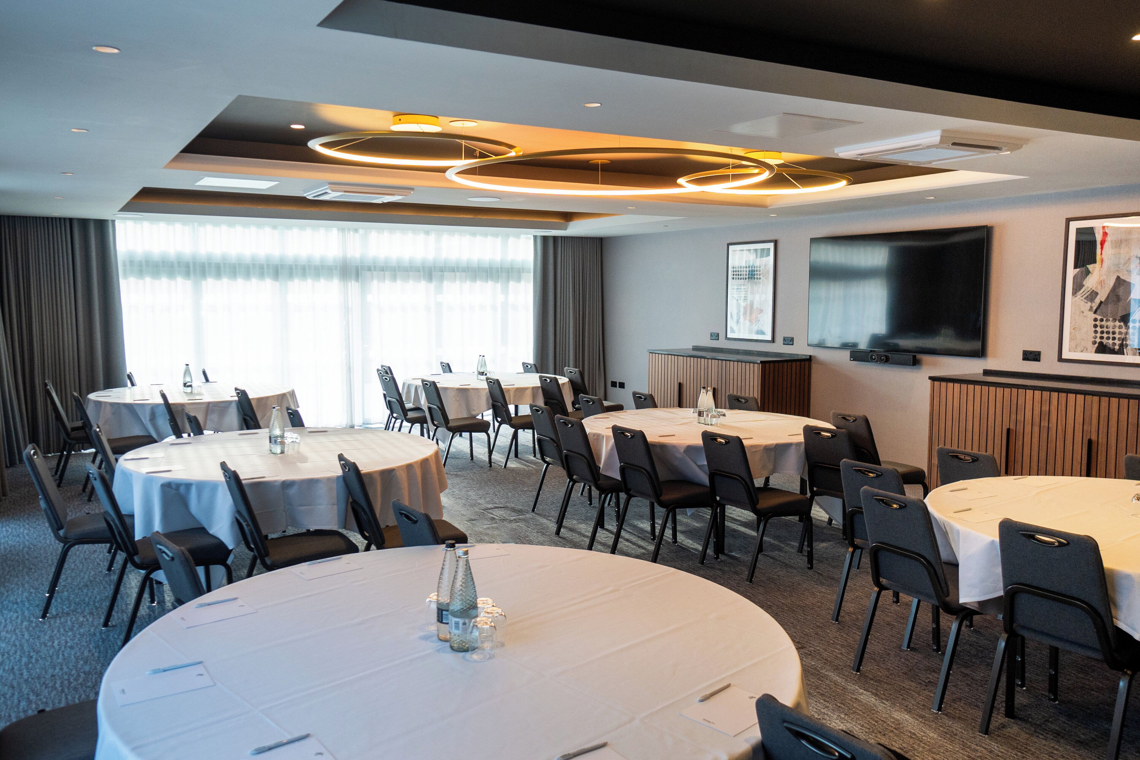 A conference room with multiple round tables, chairs, a large flat-screen TV, and a projector screen.