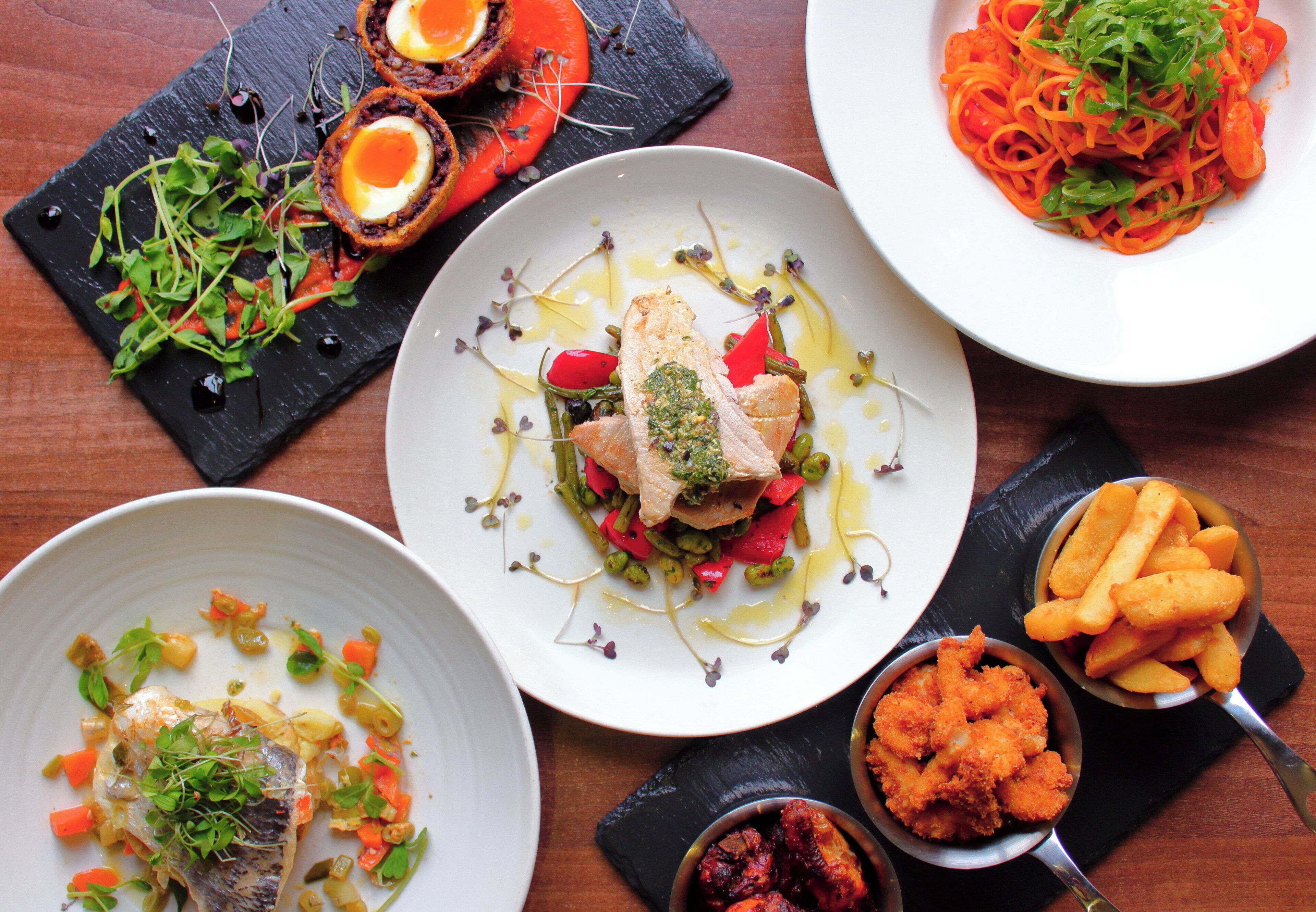 A variety of dishes on a table, including a plate with fish and vegetables, a plate with a red sauce, and a plate with a green salad.