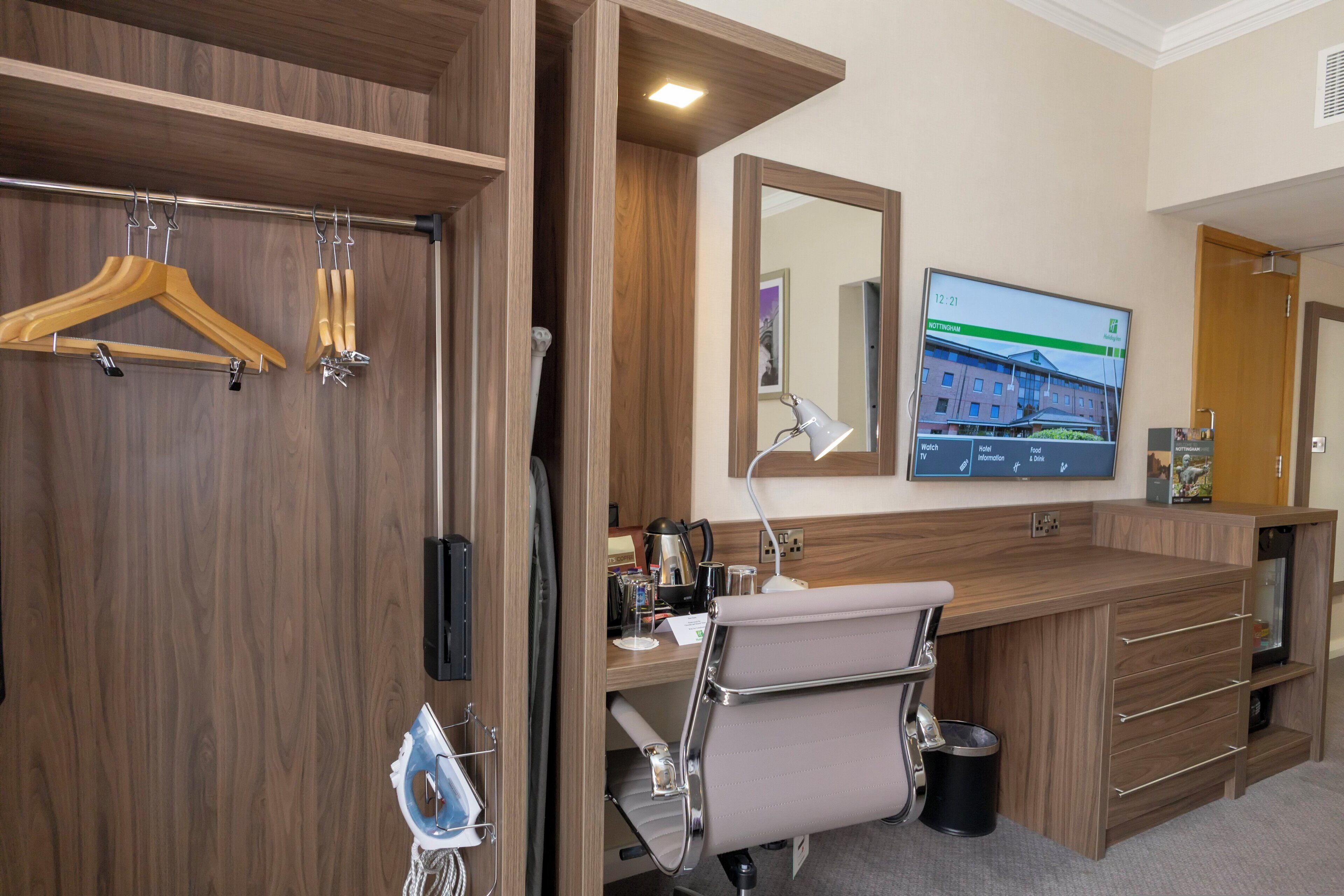 Quarto de hotel com guarda-roupa de madeira, escrivaninha com cadeira, espelho, televisão e lixeira pequena.
