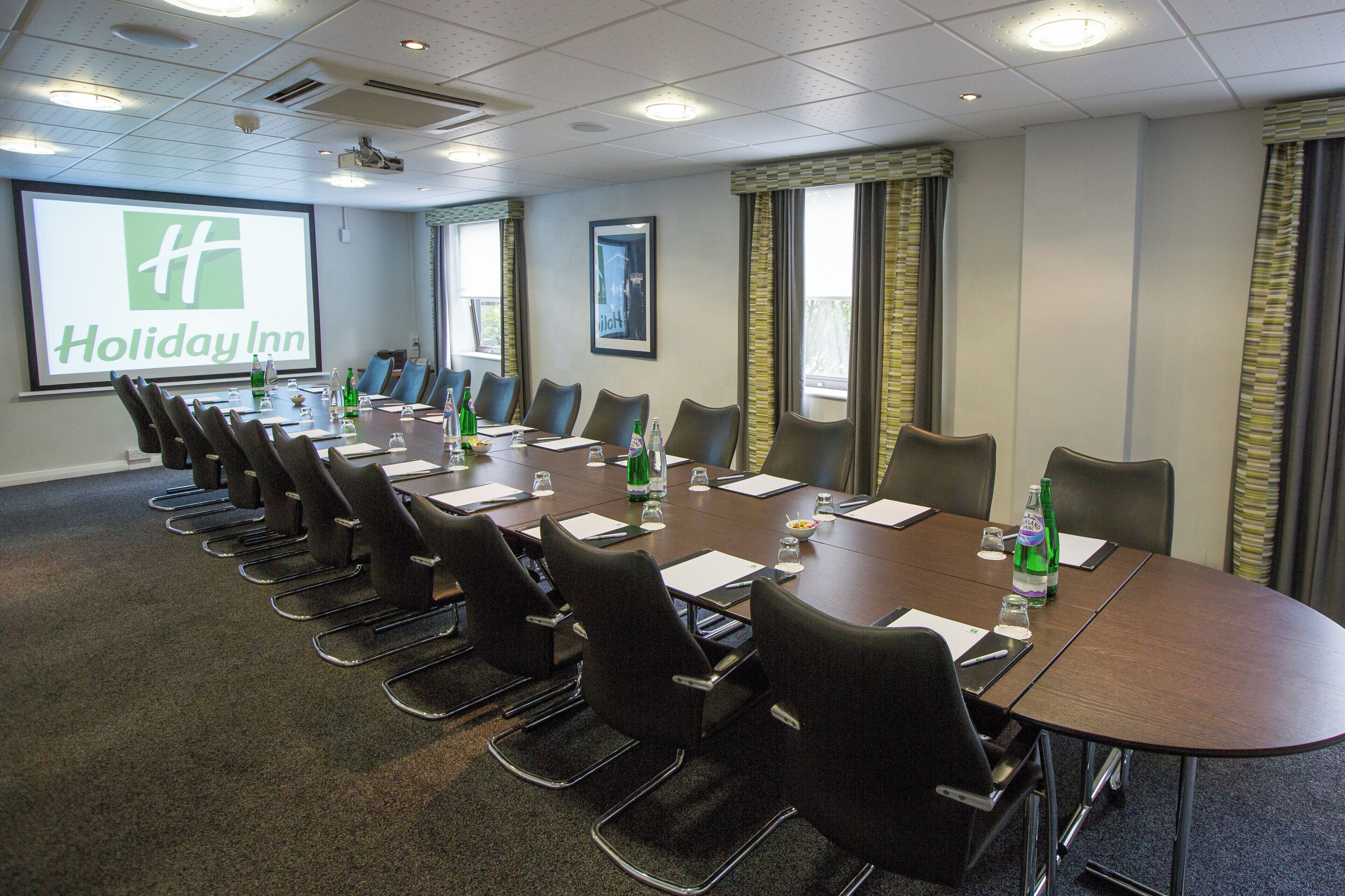 Une salle de conférence avec une longue table, des chaises et un écran de projection sur lequel est affiché le nom « Holiday Inn ».