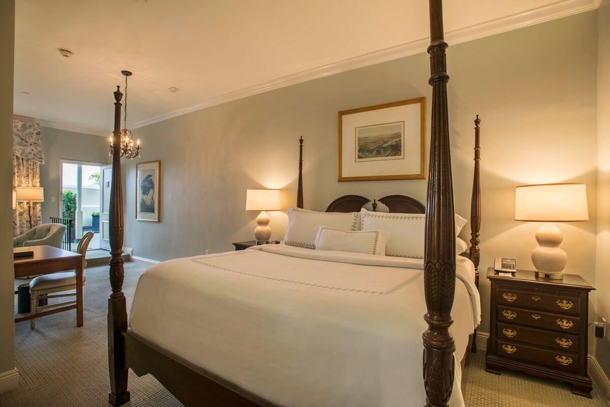 A four-poster bed with a wooden headboard, two bedside tables with lamps, and a chair in a hotel room.