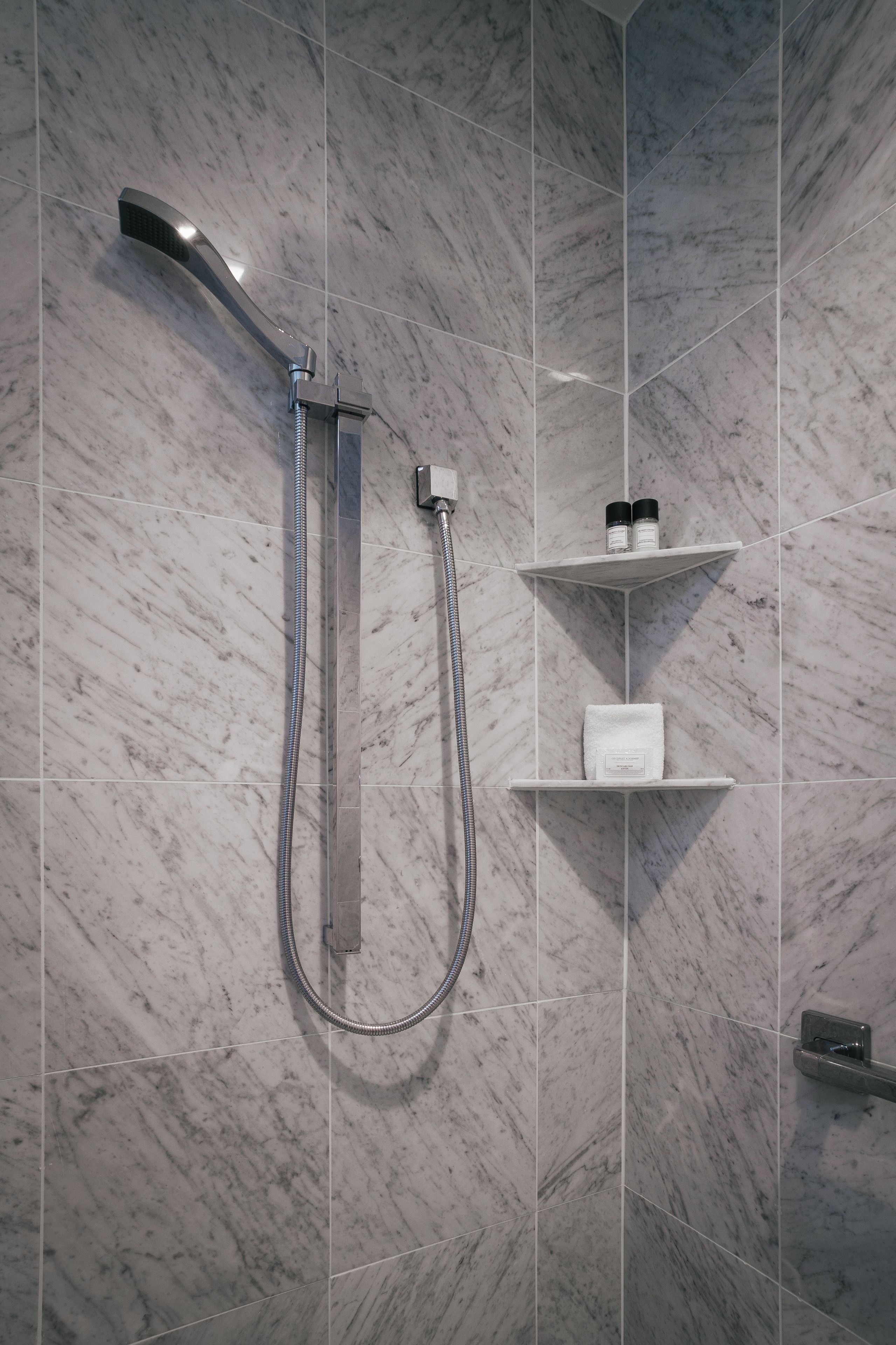 Un baño moderno con ducha, azulejos de mármol y estantes empotrados con artículos de aseo.