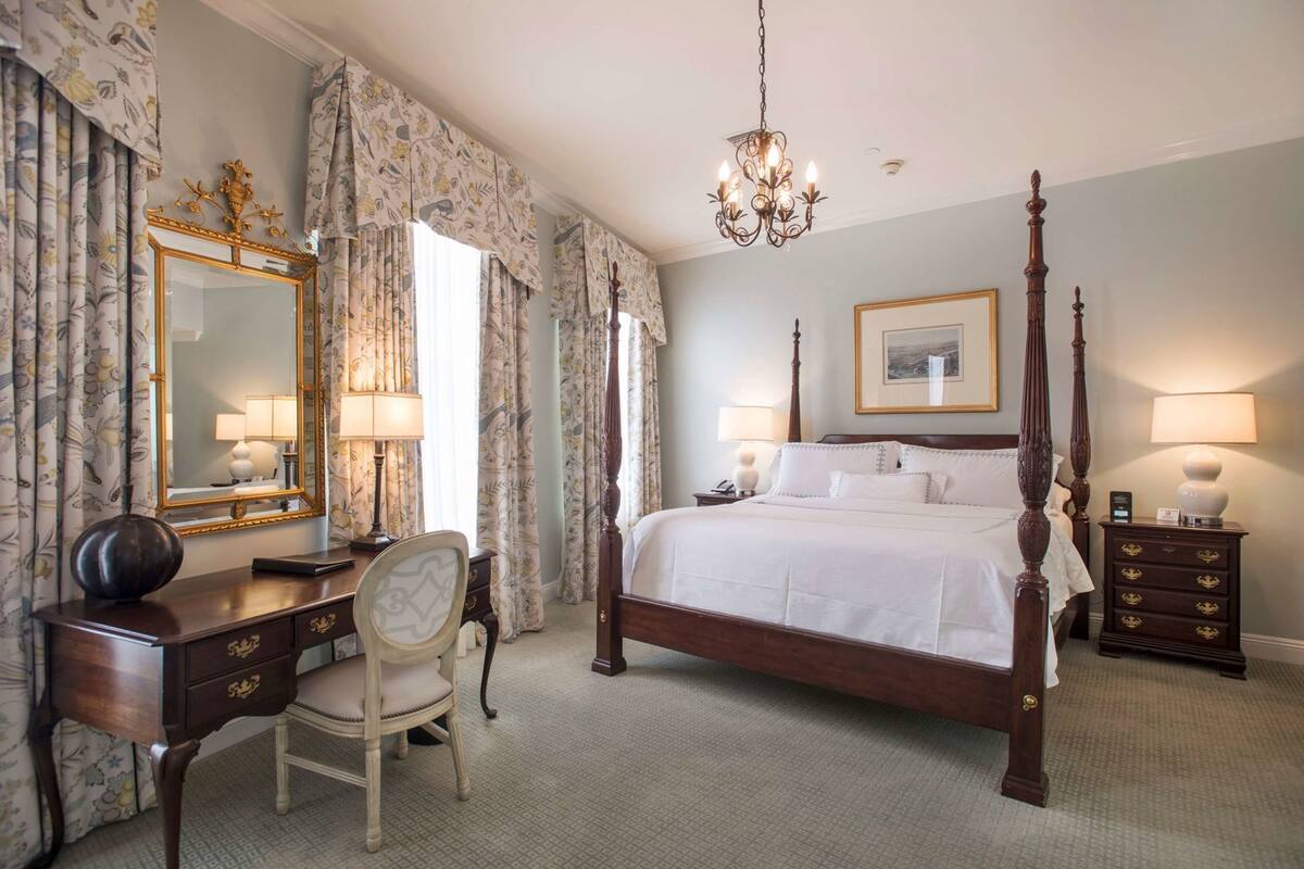 A bedroom with a four-poster bed, a desk with a chair, a large mirror, and a chandelier.