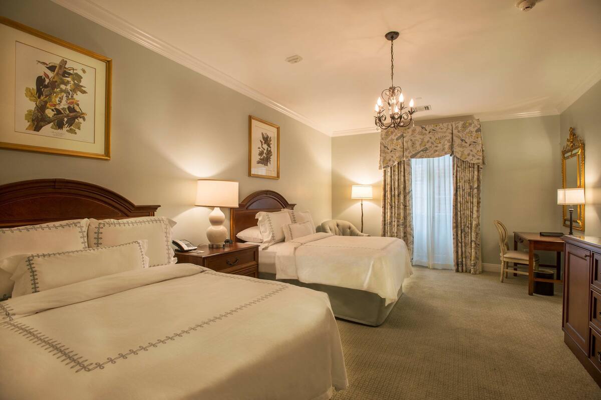 A hotel room with two beds, a chandelier, a desk with a chair, and a large window with curtains.