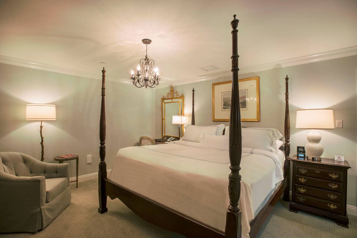 A four-poster bed with a chandelier, a chair, a bedside table, and a dresser.