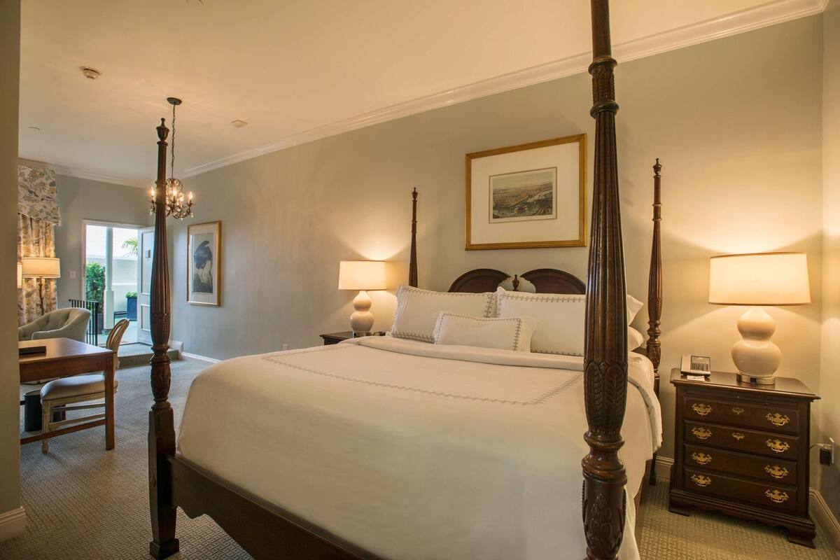 A four-poster bed with a headboard, two bedside tables with lamps, a chair, and a desk with a lamp.