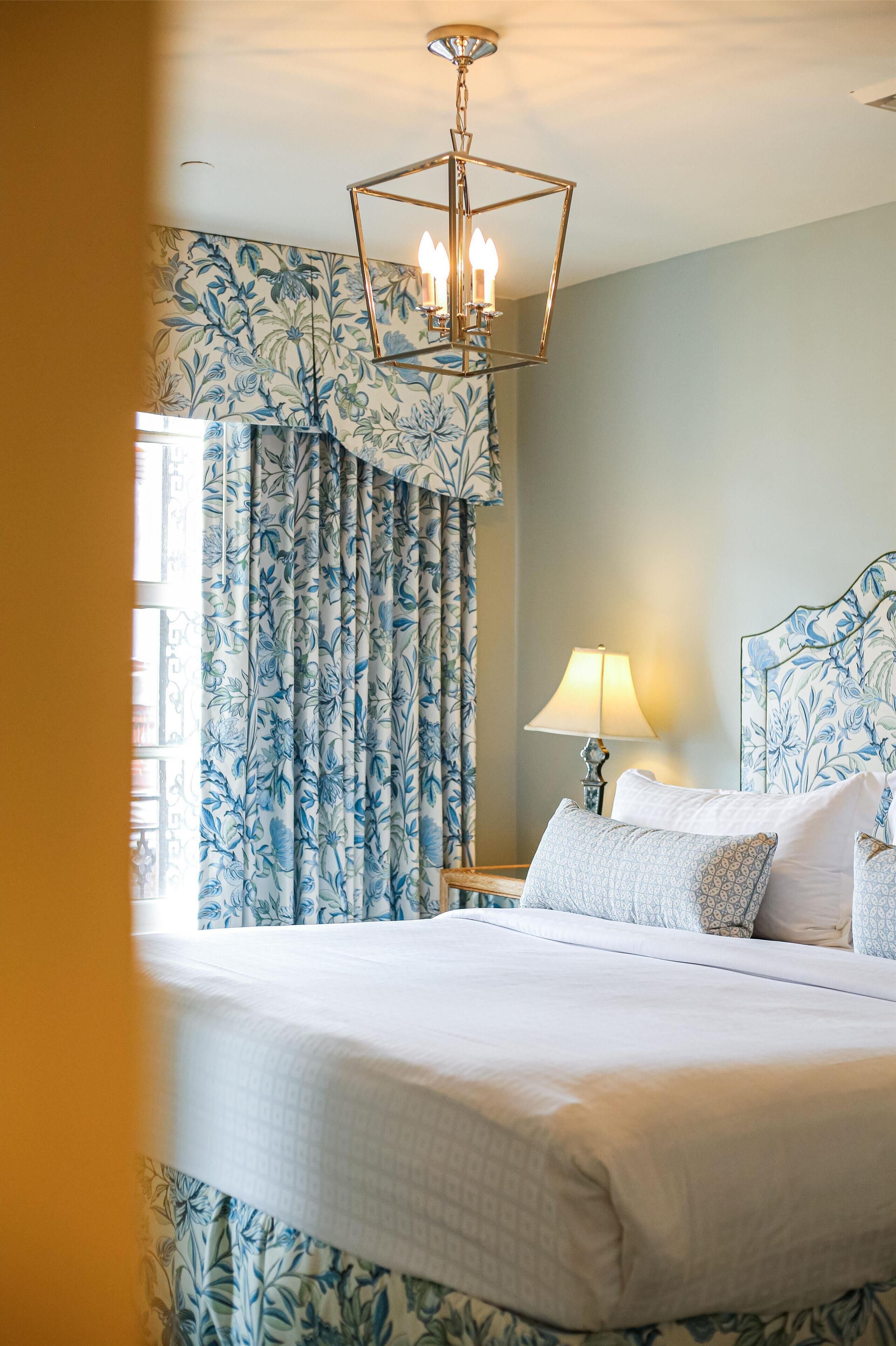 Quarto de hotel com cama, cortinas, abajur e lustre.