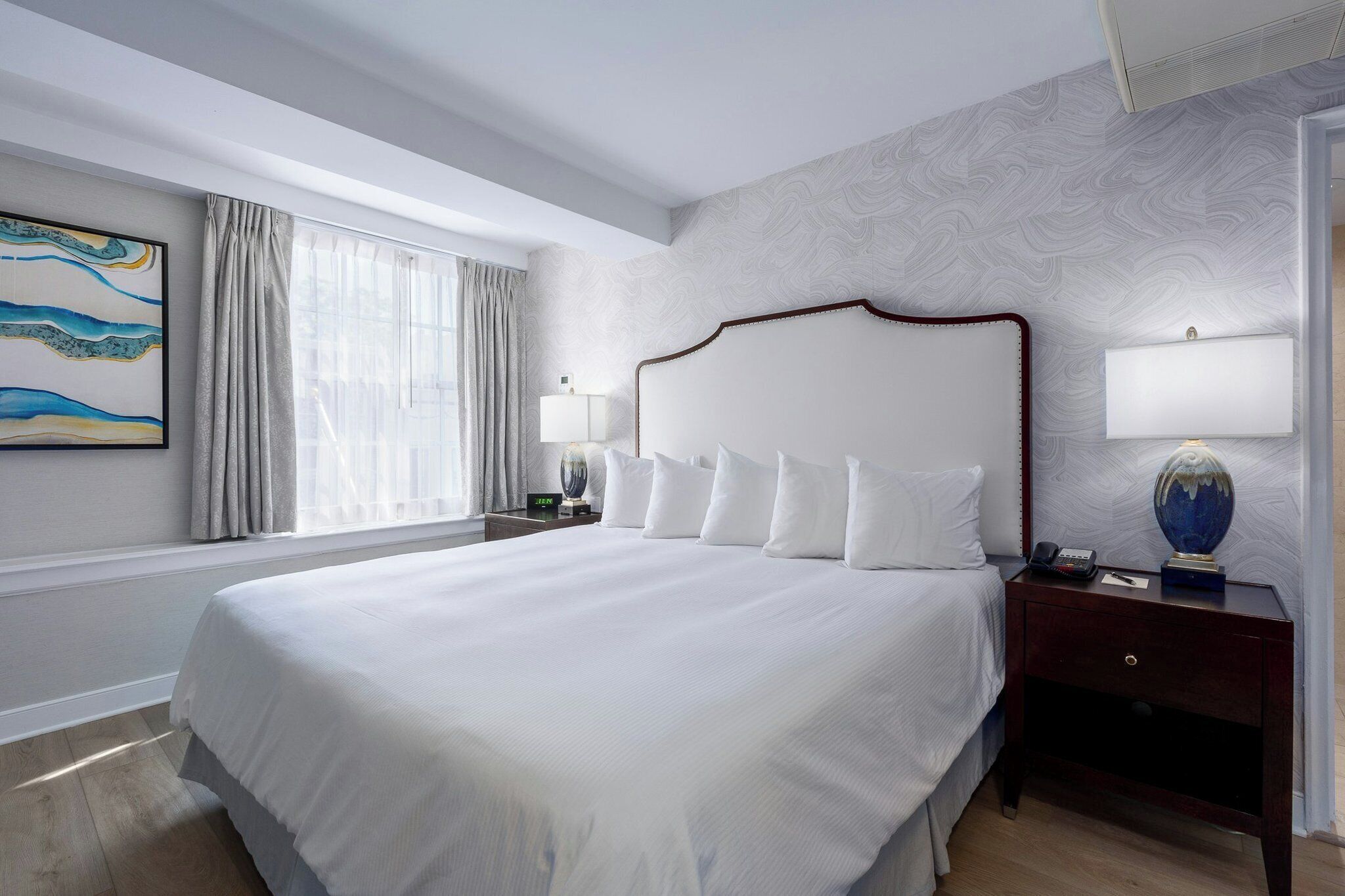A hotel room with a large bed, a nightstand with a lamp, a painting on the wall, and a window with curtains.