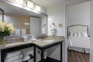 A hotel bathroom with a sink, mirror, towel rack, and a bed visible through an open door.