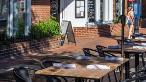 An outdoor seating area with tables and chairs, a brick building in the background, and a person walking by. - The Georgetown Inn (Washington)