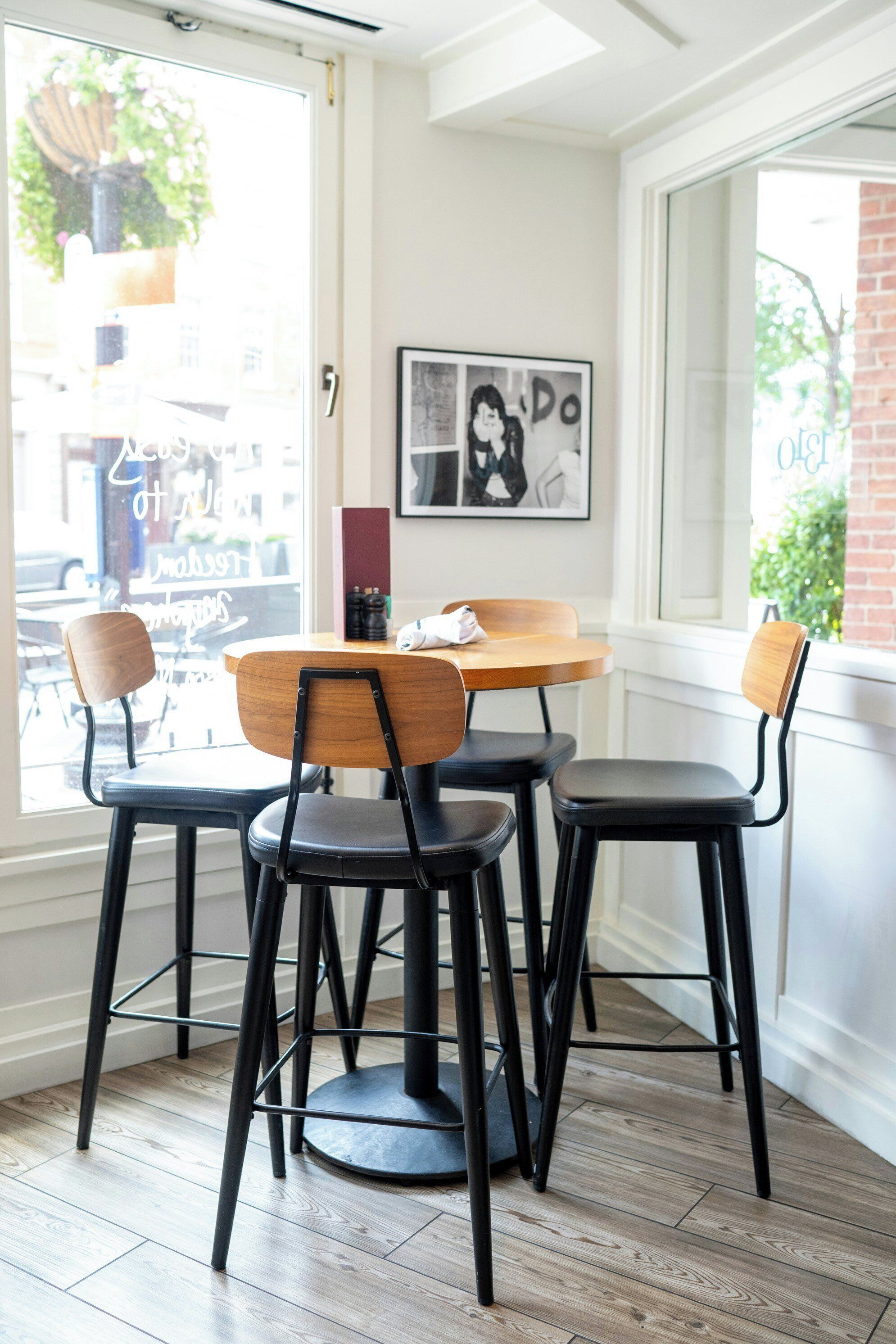 A corner seating area with a round table and four chairs, a framed picture on the wall, and a window with a view of the street.