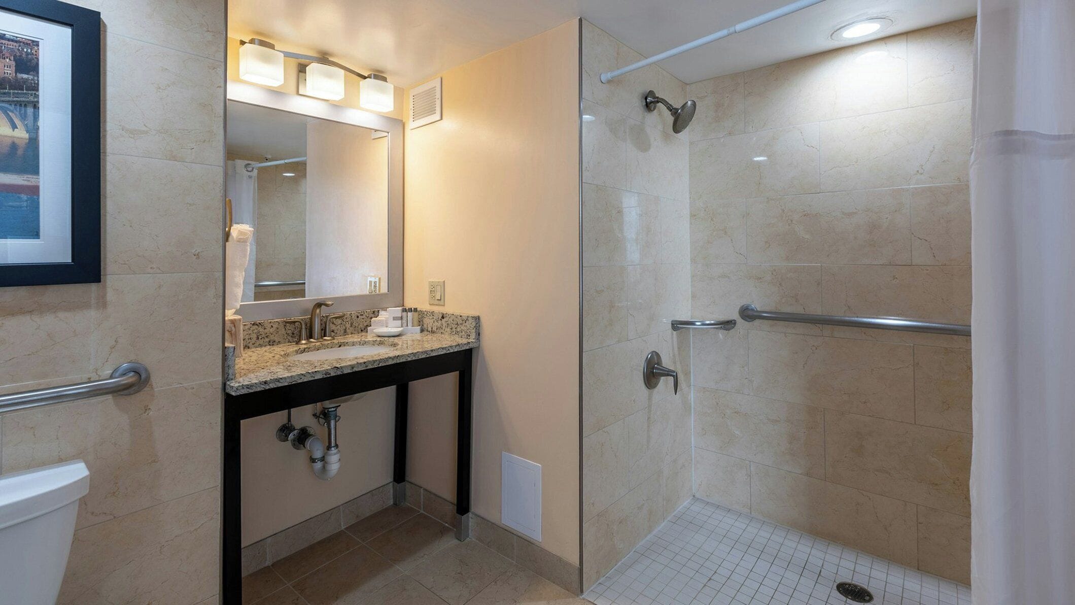 A bathroom with a granite countertop, a large mirror, a shower with a glass door, and a handrail.